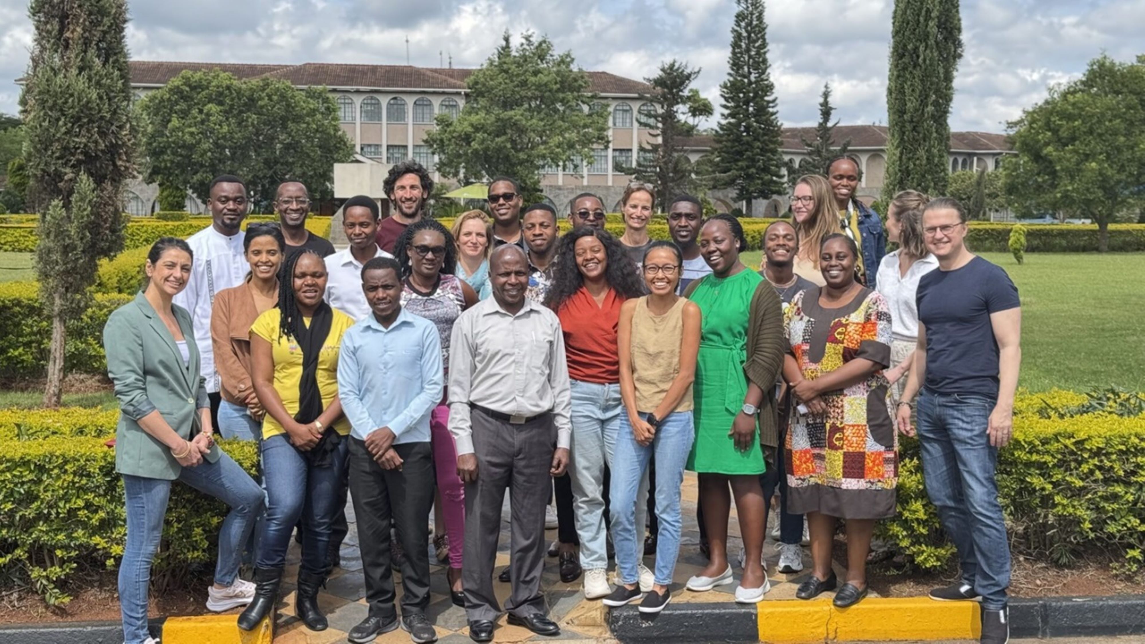 A diverse group of individuals stands together outdoors, framed by lush greenery and a historic building in the background. They wear a variety of colorful clothing, showcasing personal styles. The scene is bright, with a partly cloudy sky, suggesting a warm day. The atmosphere feels friendly and collaborative.