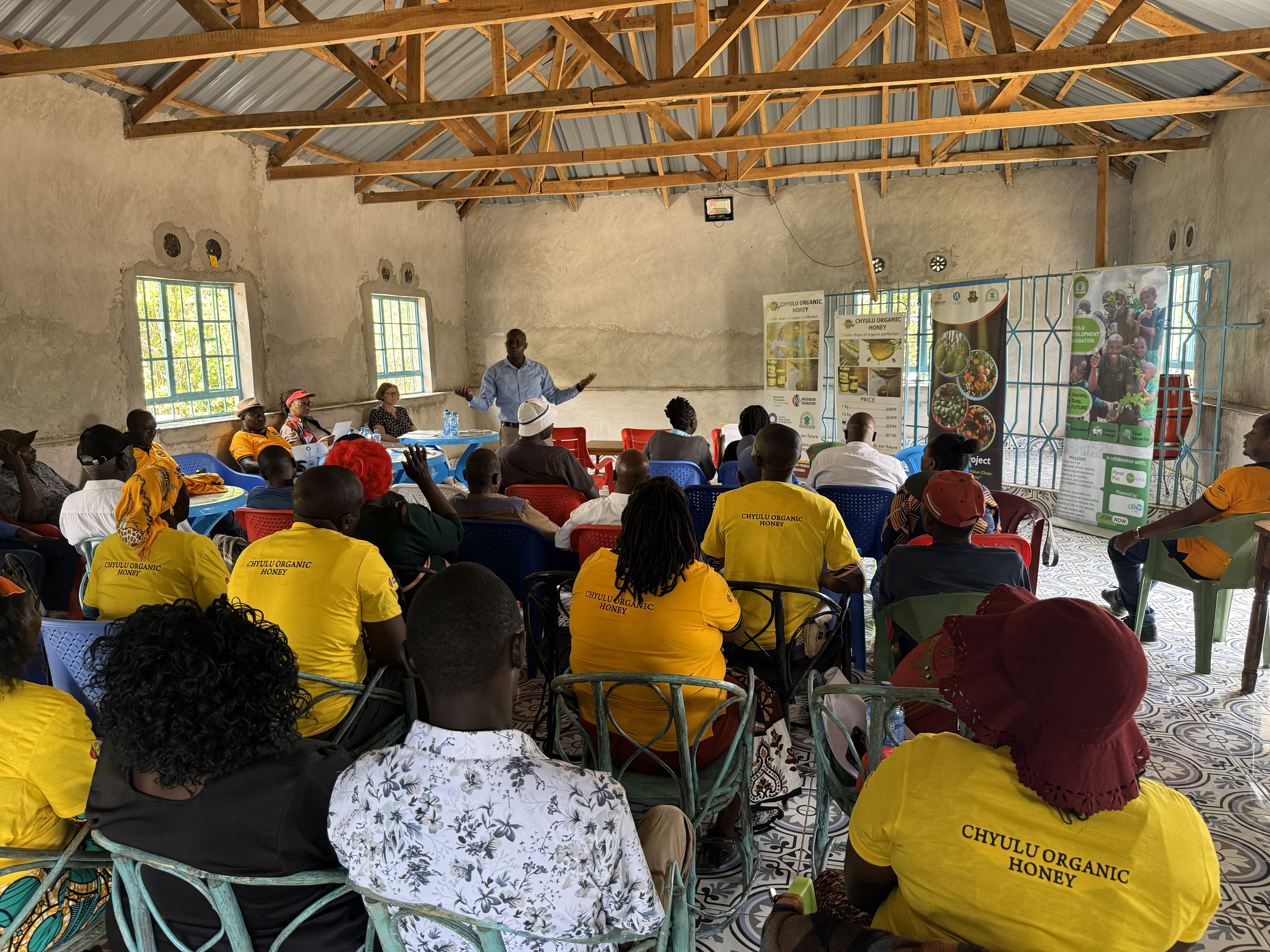 A group of people seated in a room, listening to a speaker, with banners about organic honey in the background.