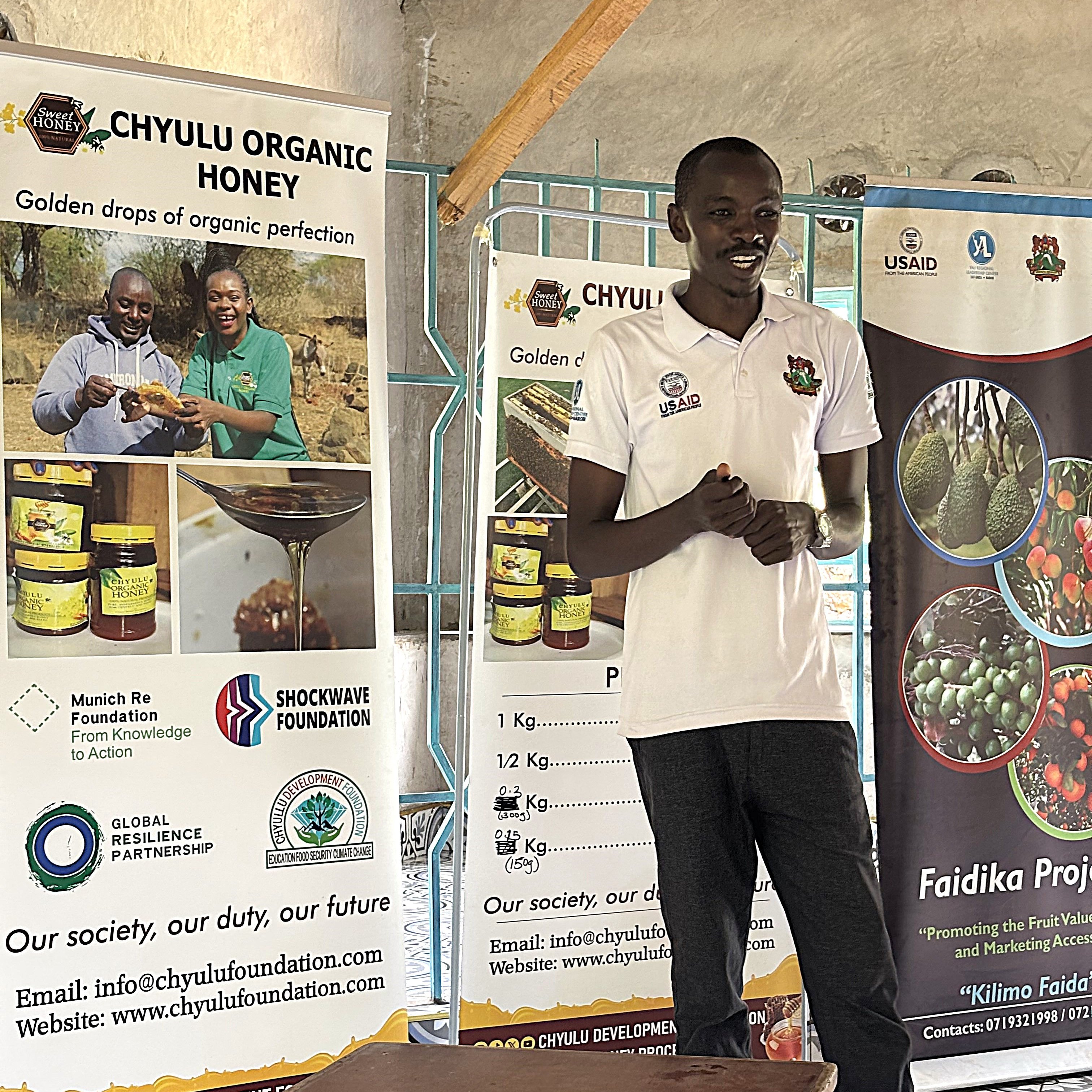 A person stands in front of banners promoting Chyulu Organic Honey and related projects in a community setting.