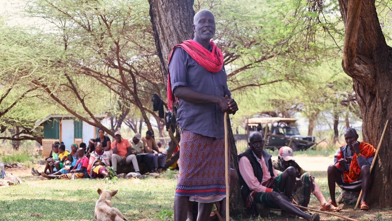 A group of people sitting under trees, with one person standing holding a stick, and a dog lying on the grass.
