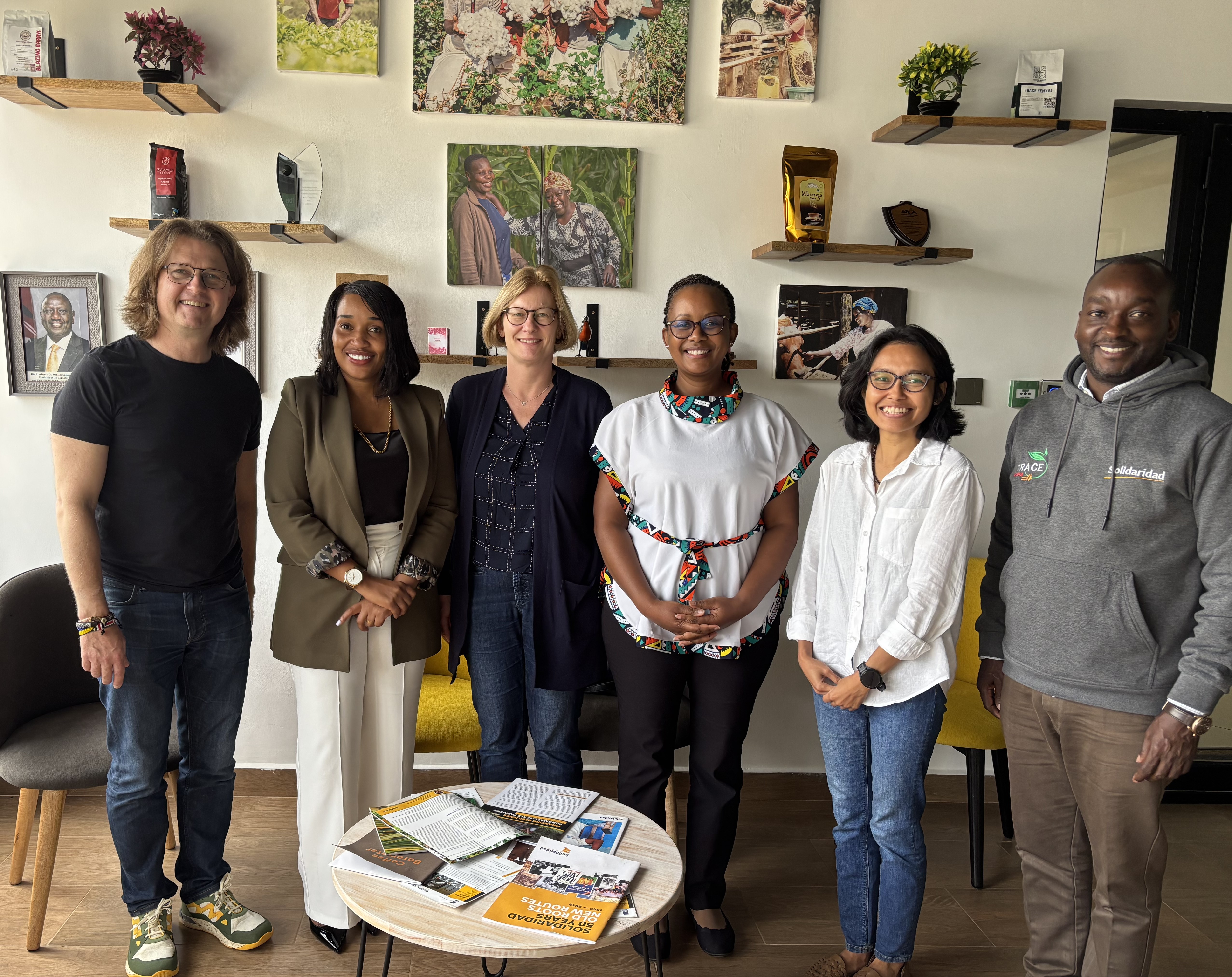 A group of six people stands together in a room with wall art and shelves displaying items.