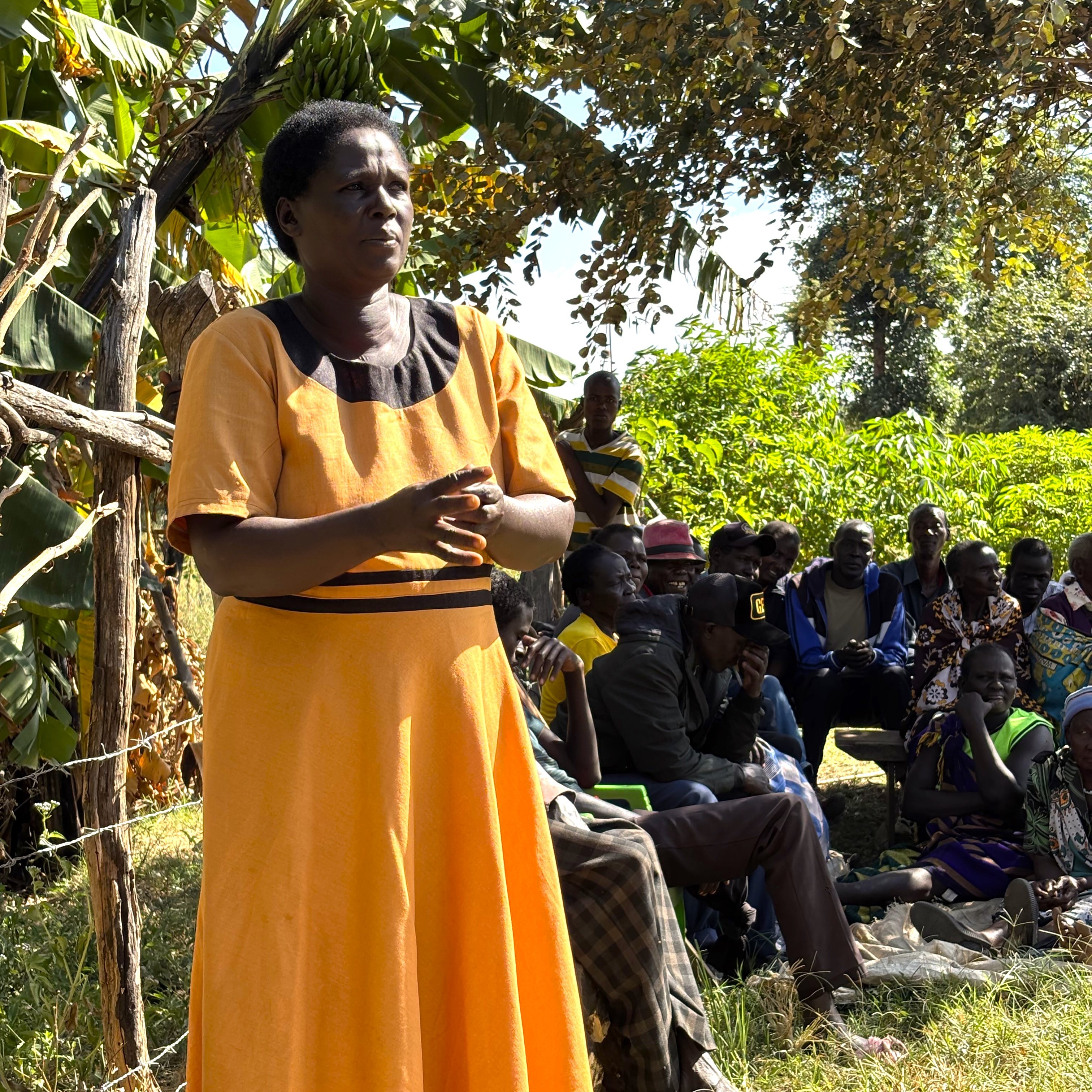 A woman in an orange dress stands speaking to a group seated outdoors among trees and greenery.