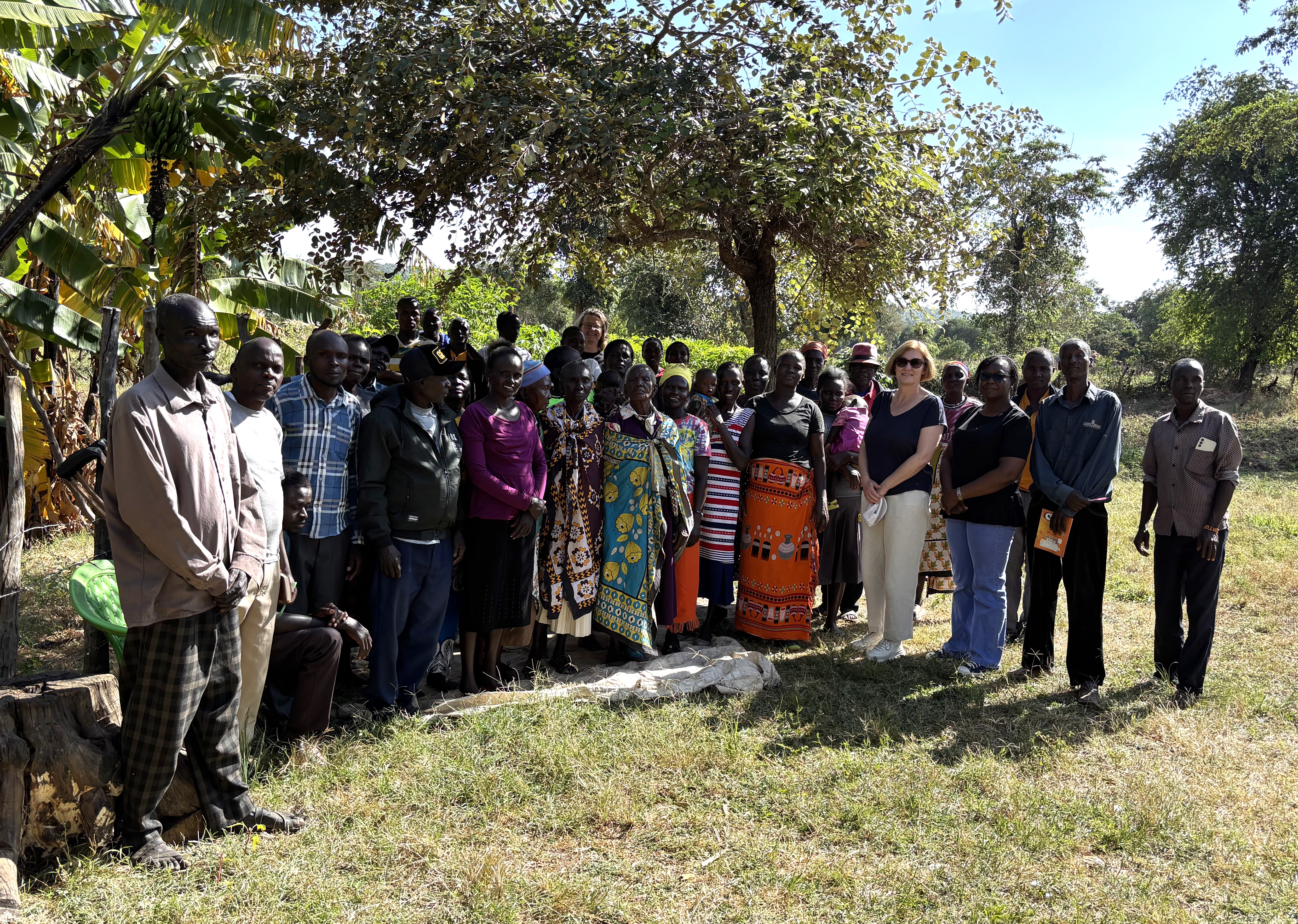 A group of five people stands outdoors in a grassy area with trees and a wooden fence in the background.