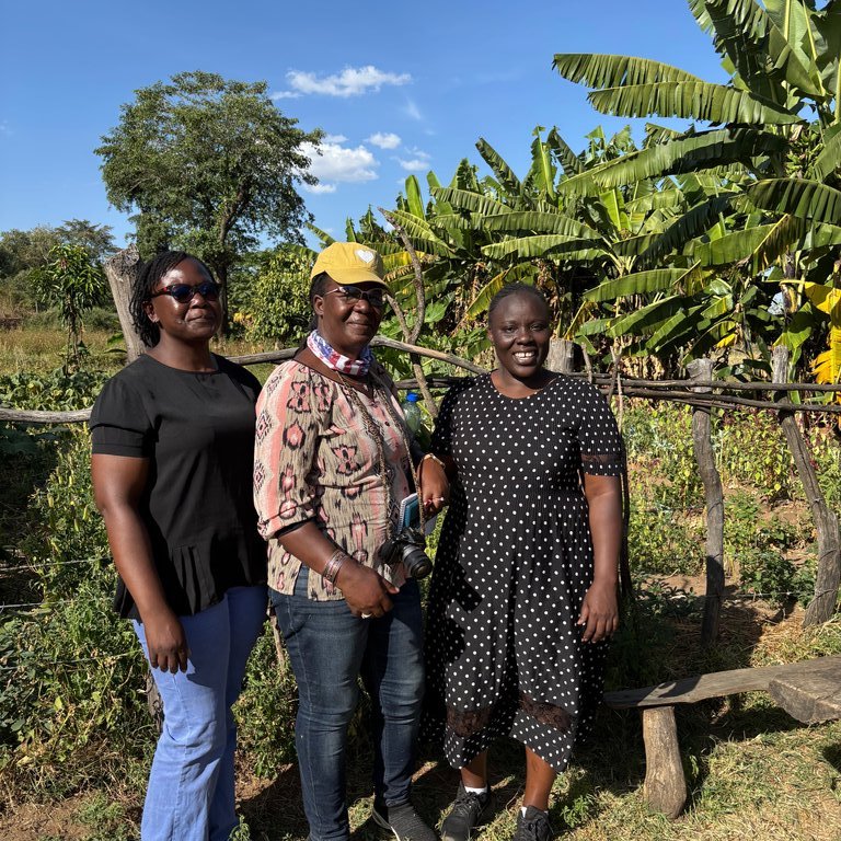 A group of five people stands outdoors in a grassy area with trees and a wooden fence in the background.
