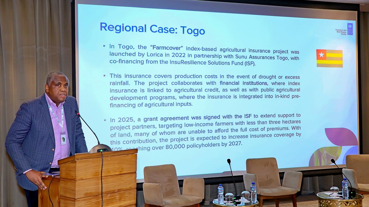 A speaker presents information on agricultural insurance in Togo, with a projector screen displaying details.