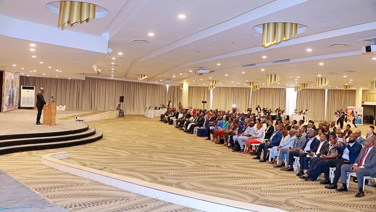 A speaker at a podium addresses an audience seated in a conference hall with curtains and decorative lighting.