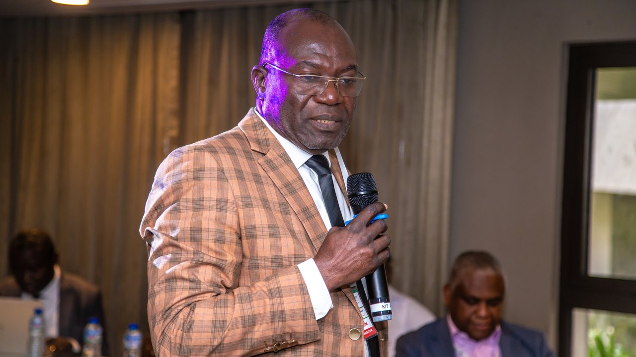 A man in a checkered suit speaks into a microphone at a conference, with blurred faces of attendees in the background.
