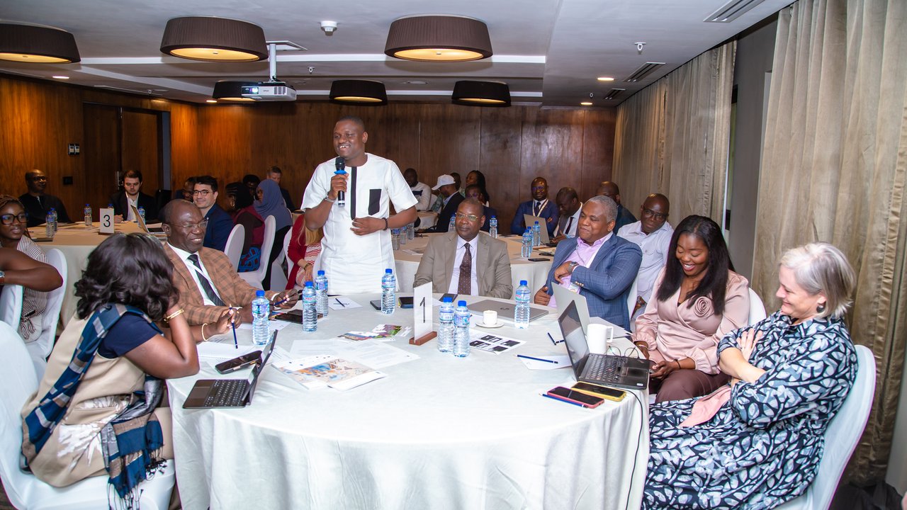 A group of people seated around a table in a conference room, some engaged in discussion and others using laptops.