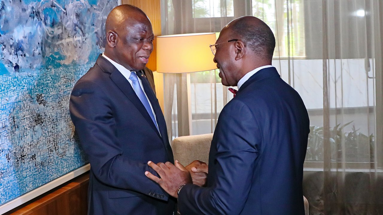 Two men in suits shake hands in a well-lit room with a large abstract painting on the wall.