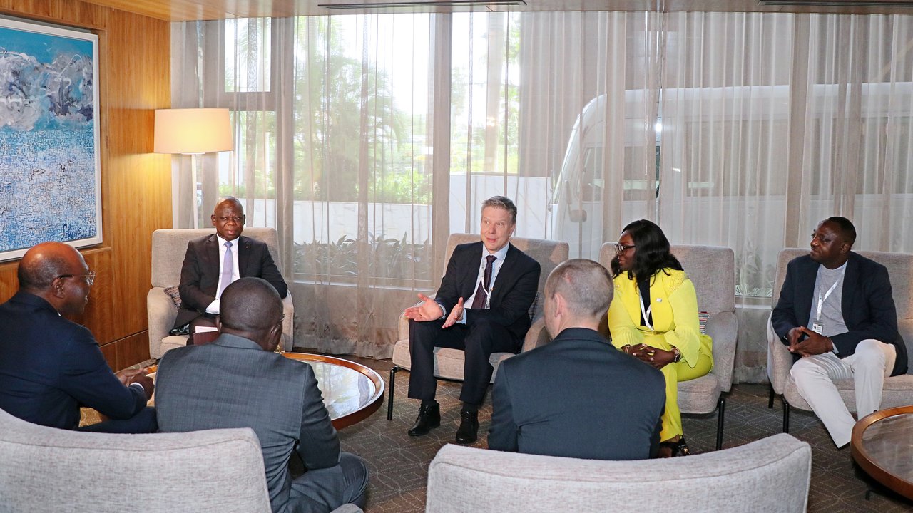 A group of six people seated in a meeting room with large windows and a painting on the wall.