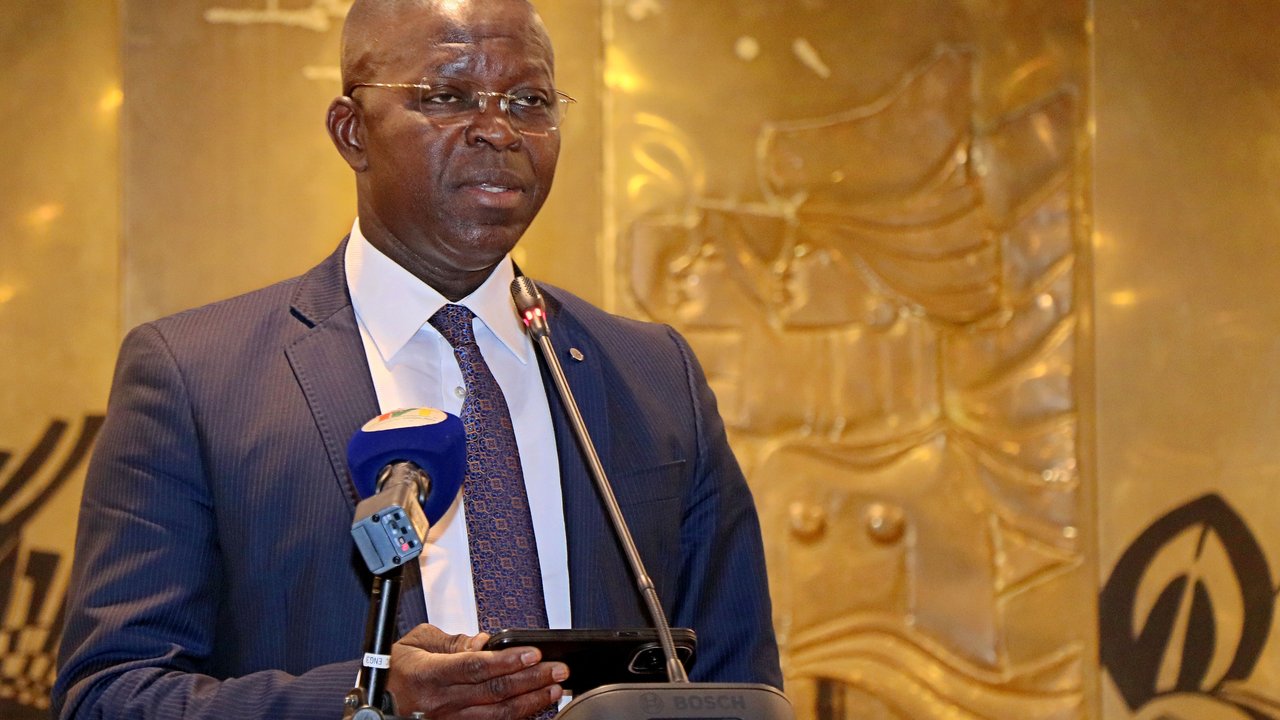 A man in a suit speaks at a podium with a microphone, set against a decorative gold background.