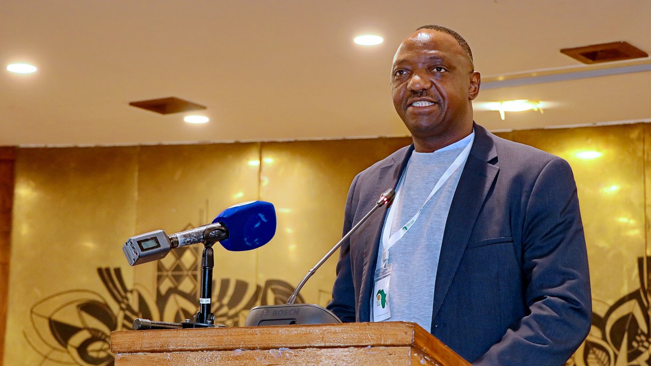 A speaker at a podium with microphones, wearing a blazer and a badge, in a conference setting.
