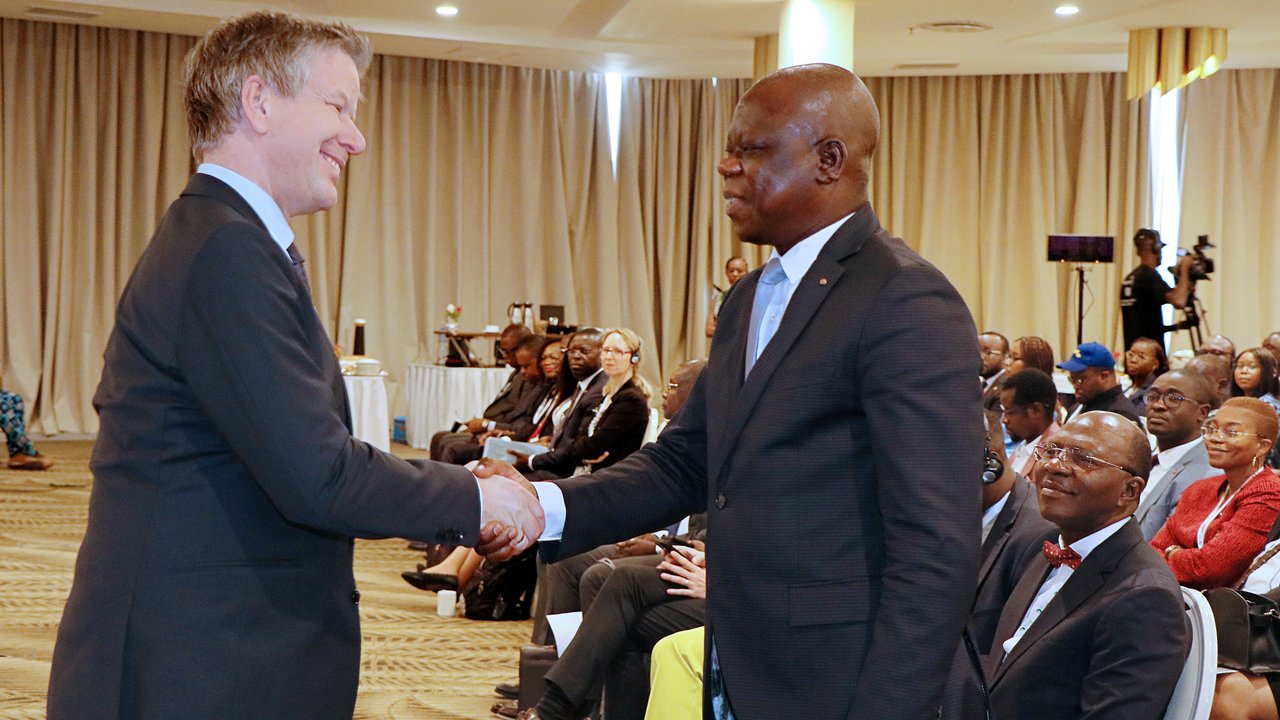 Two men in business attire shake hands in a conference setting, with an audience seated in the background.
