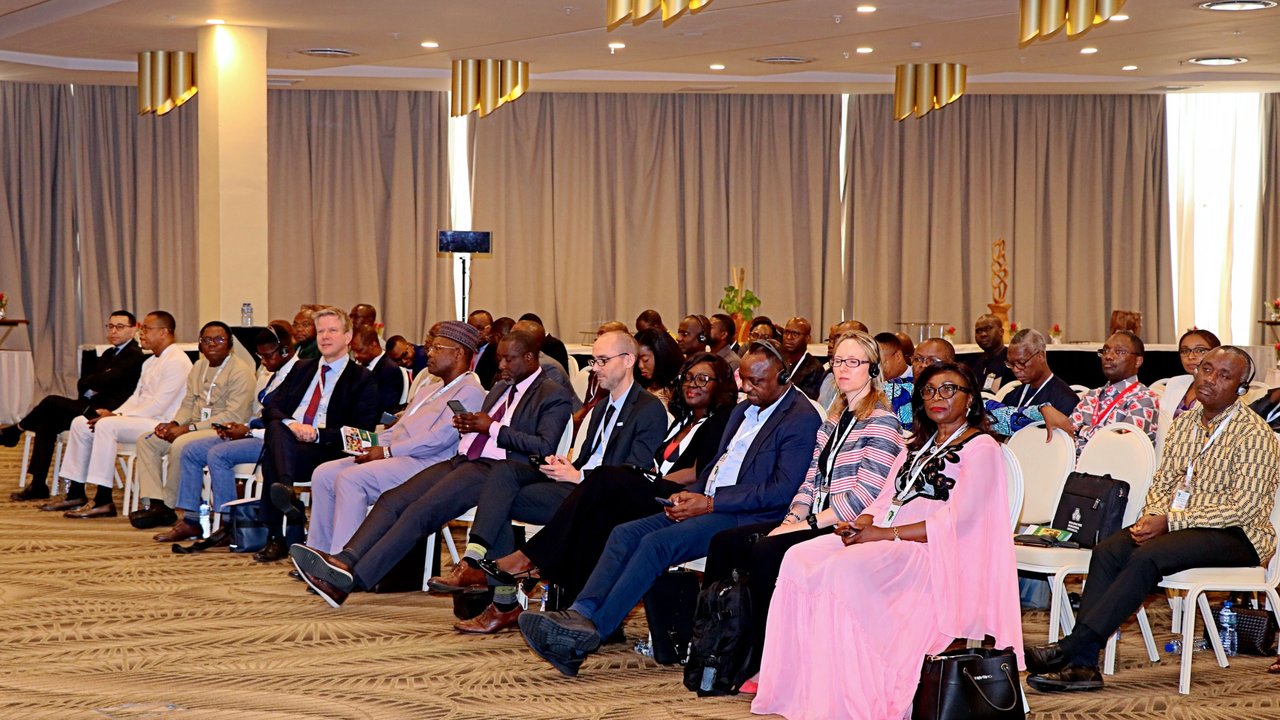 A diverse audience seated in a conference room, facing a speaker, with curtains and decorative elements in the background.