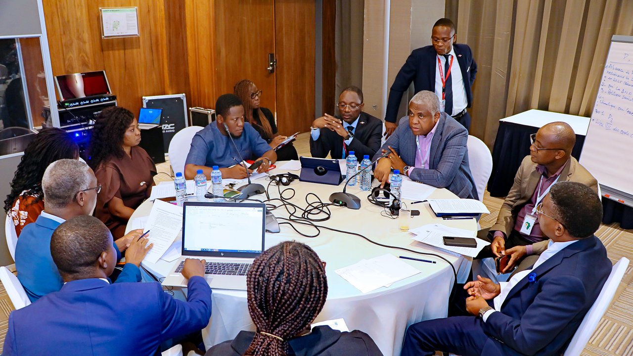 A group of people in business attire seated around a table with laptops, papers, and water bottles during a meeting.