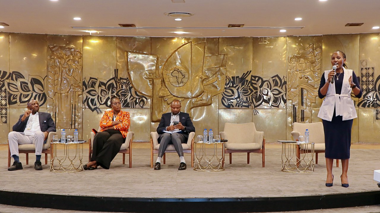 A panel discussion with four speakers on stage, seated and one standing, in front of a decorative backdrop.