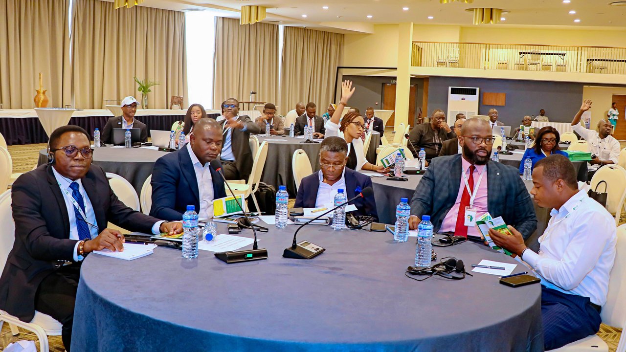 A conference room with several people seated at tables, some raising hands, and water bottles on the tables.
