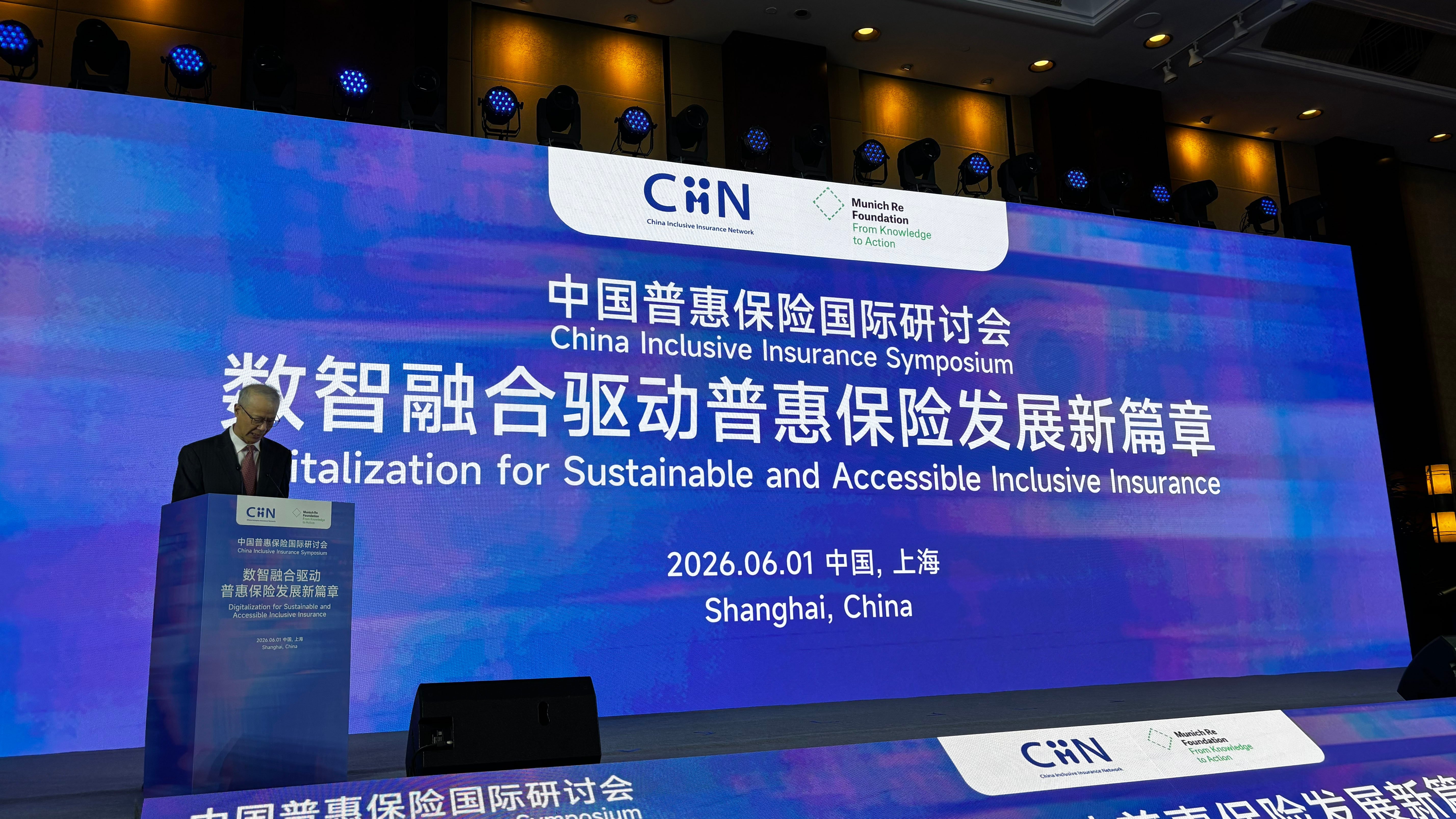 A speaker at a podium in front of a large screen displaying event details in Chinese and English.