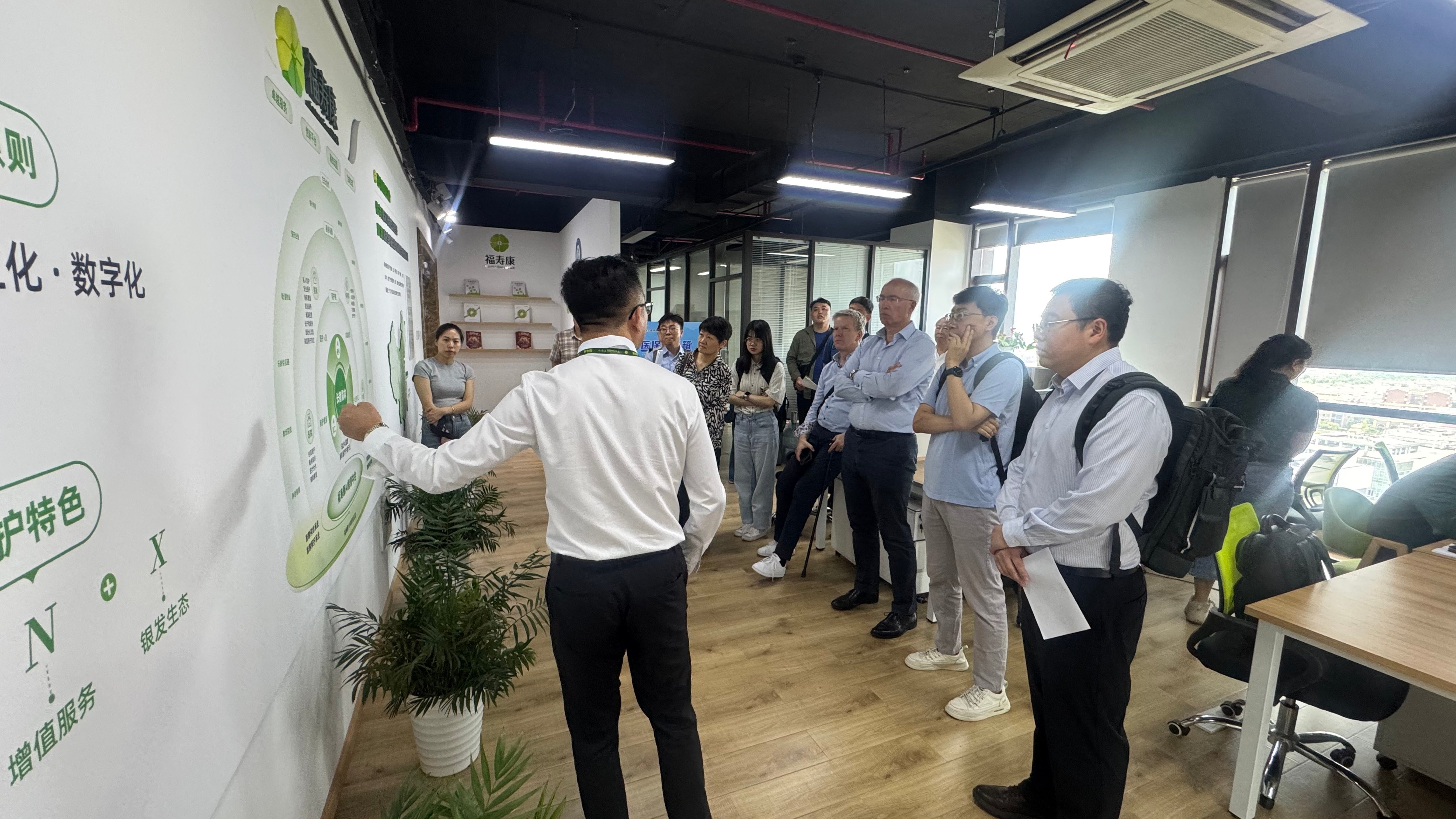 A group of people in an office listening to a presentation by a speaker in front of a wall with graphics.