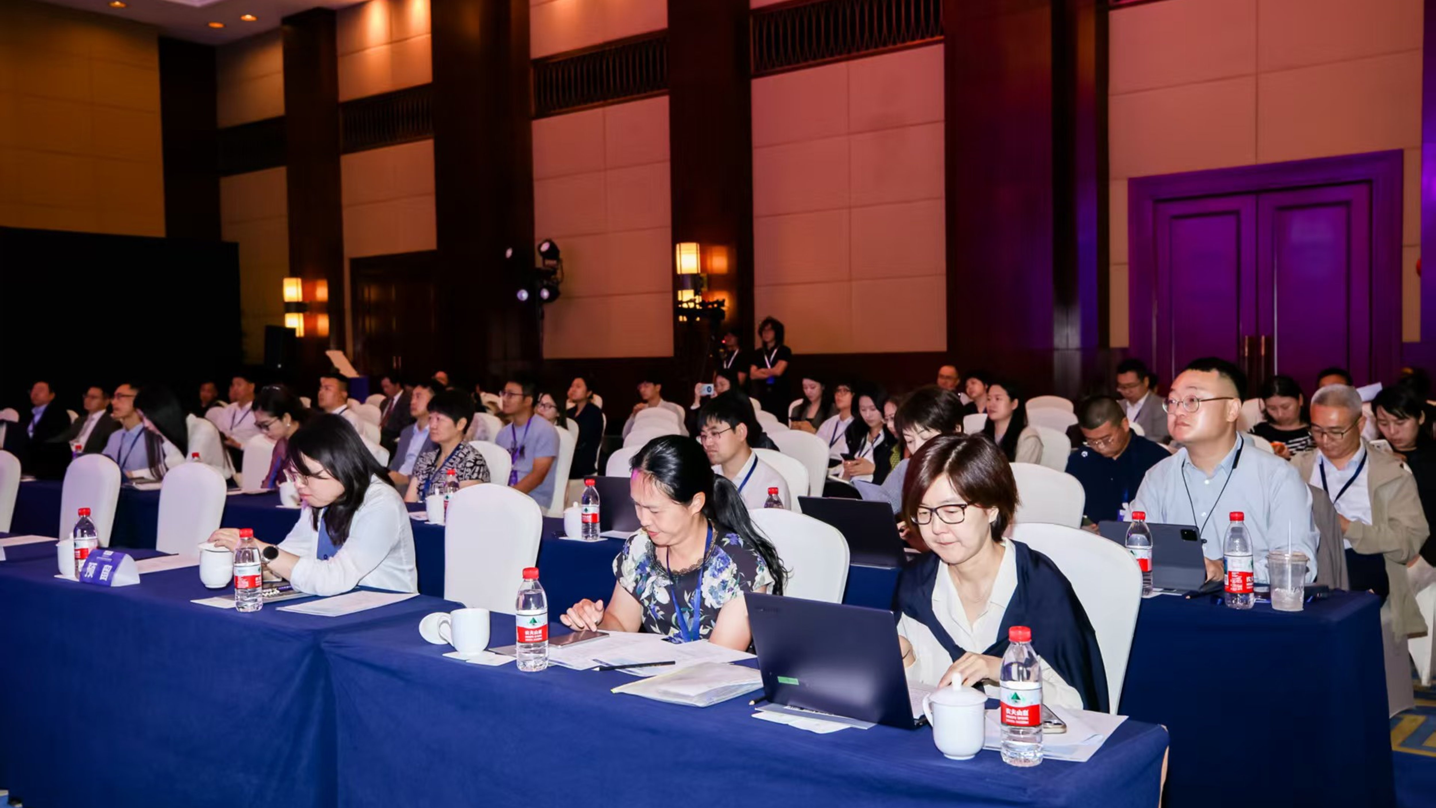 A conference room with attendees seated at tables, focused on presentations, with laptops and water bottles visible.