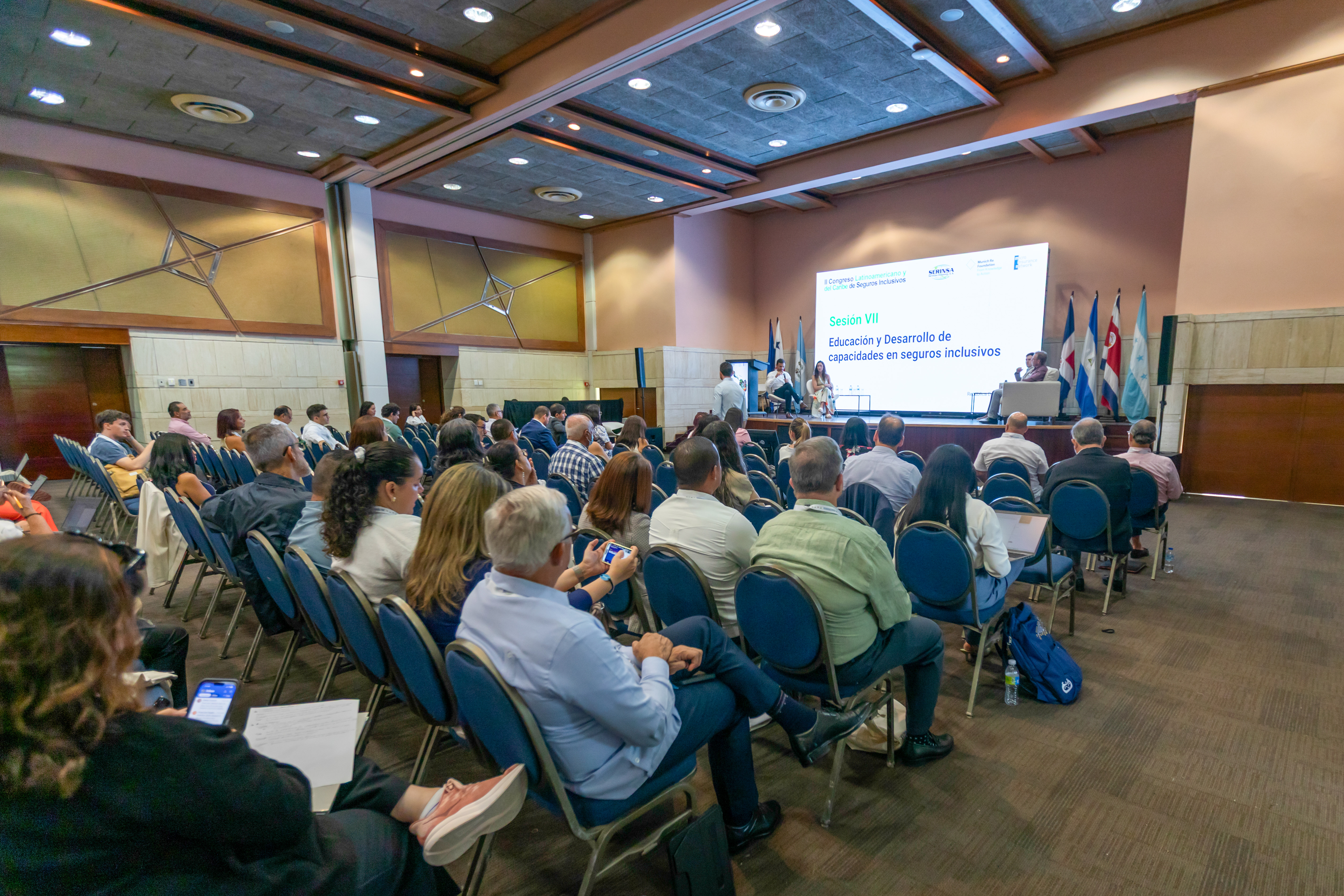 A conference room with attendees seated, a presentation screen, and flags in the background.