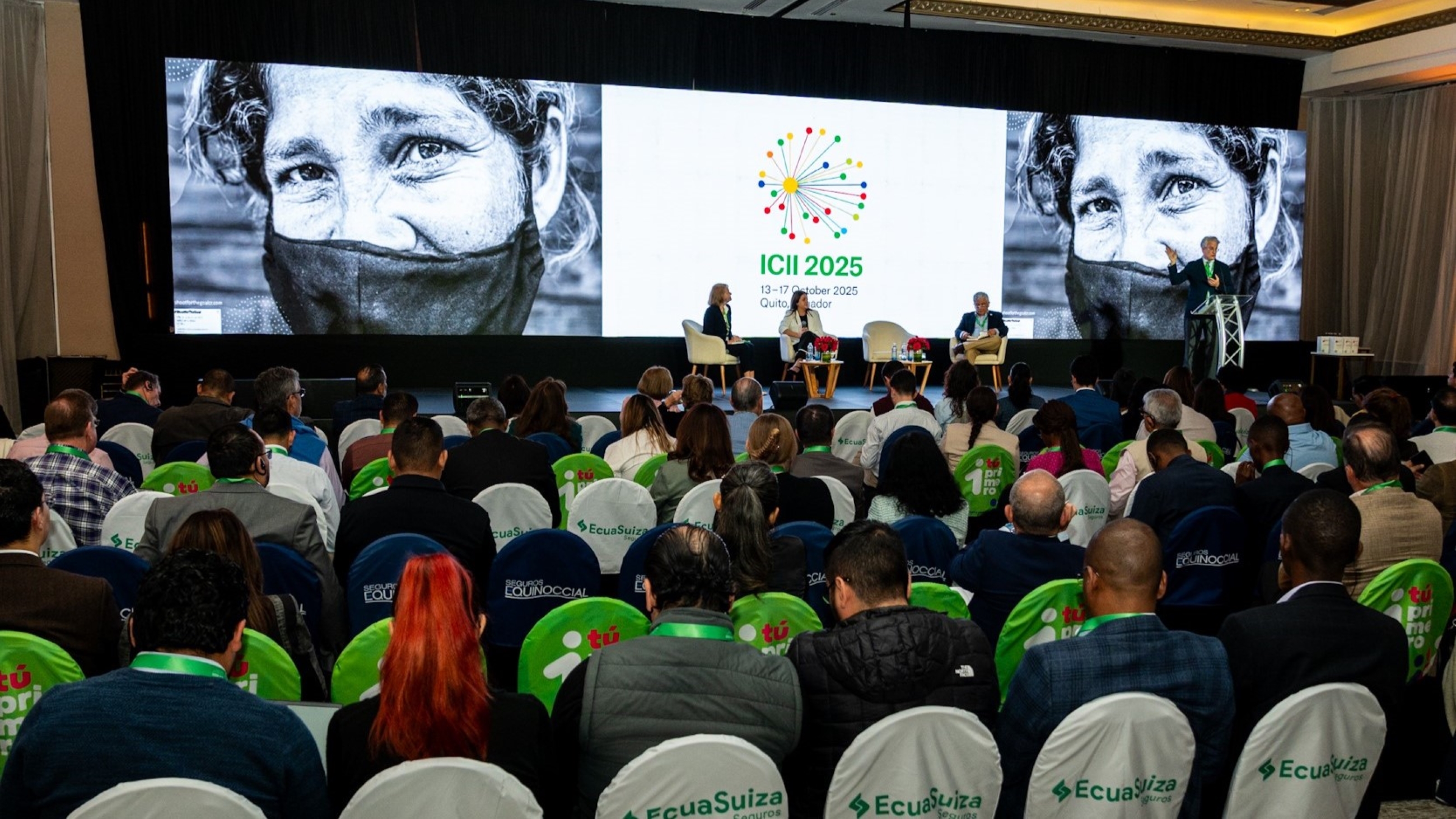 A conference scene with attendees seated, a speaker panel, and a large screen displaying event details.
