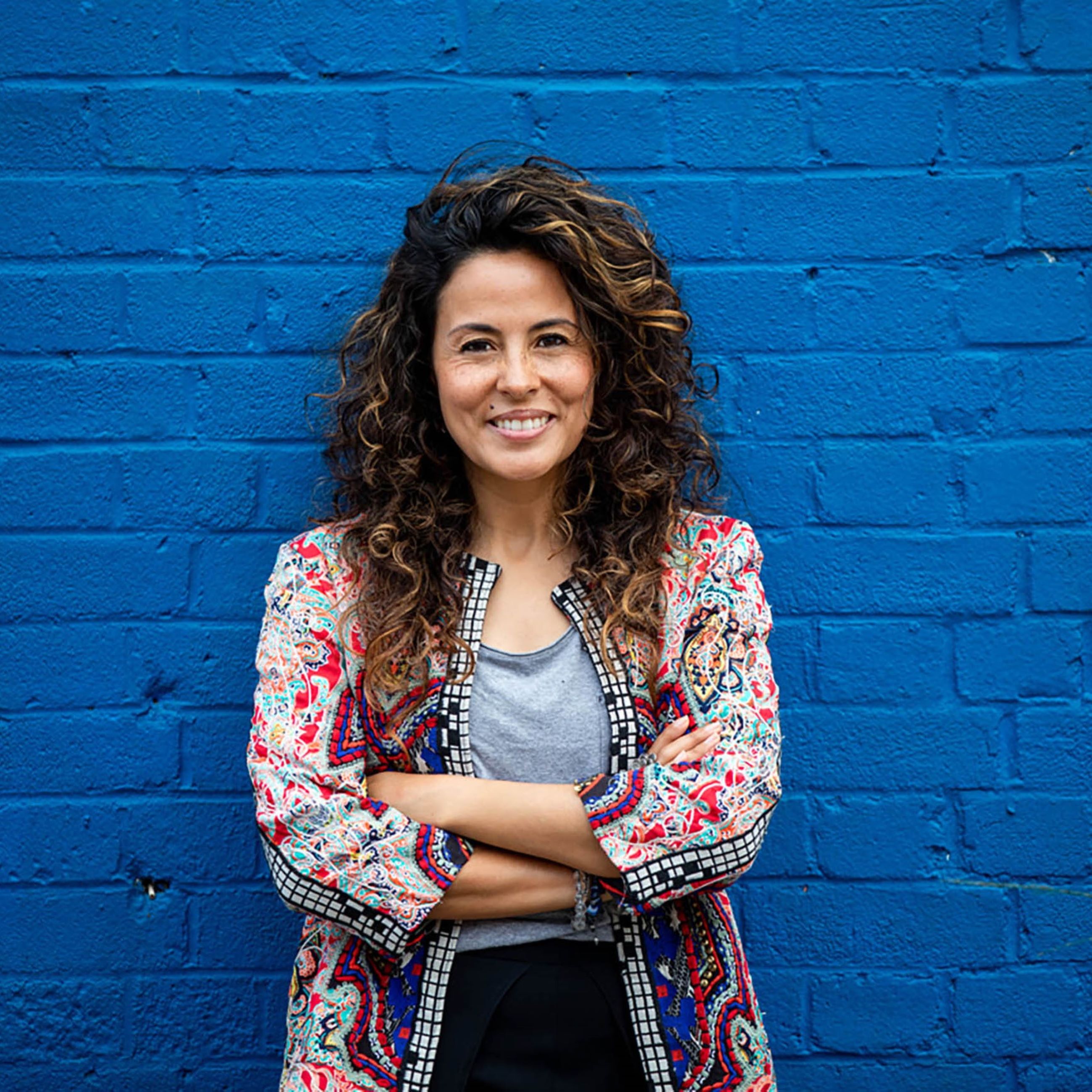 A person with curly hair stands against a bright blue brick wall, wearing a colorful patterned jacket and gray top.