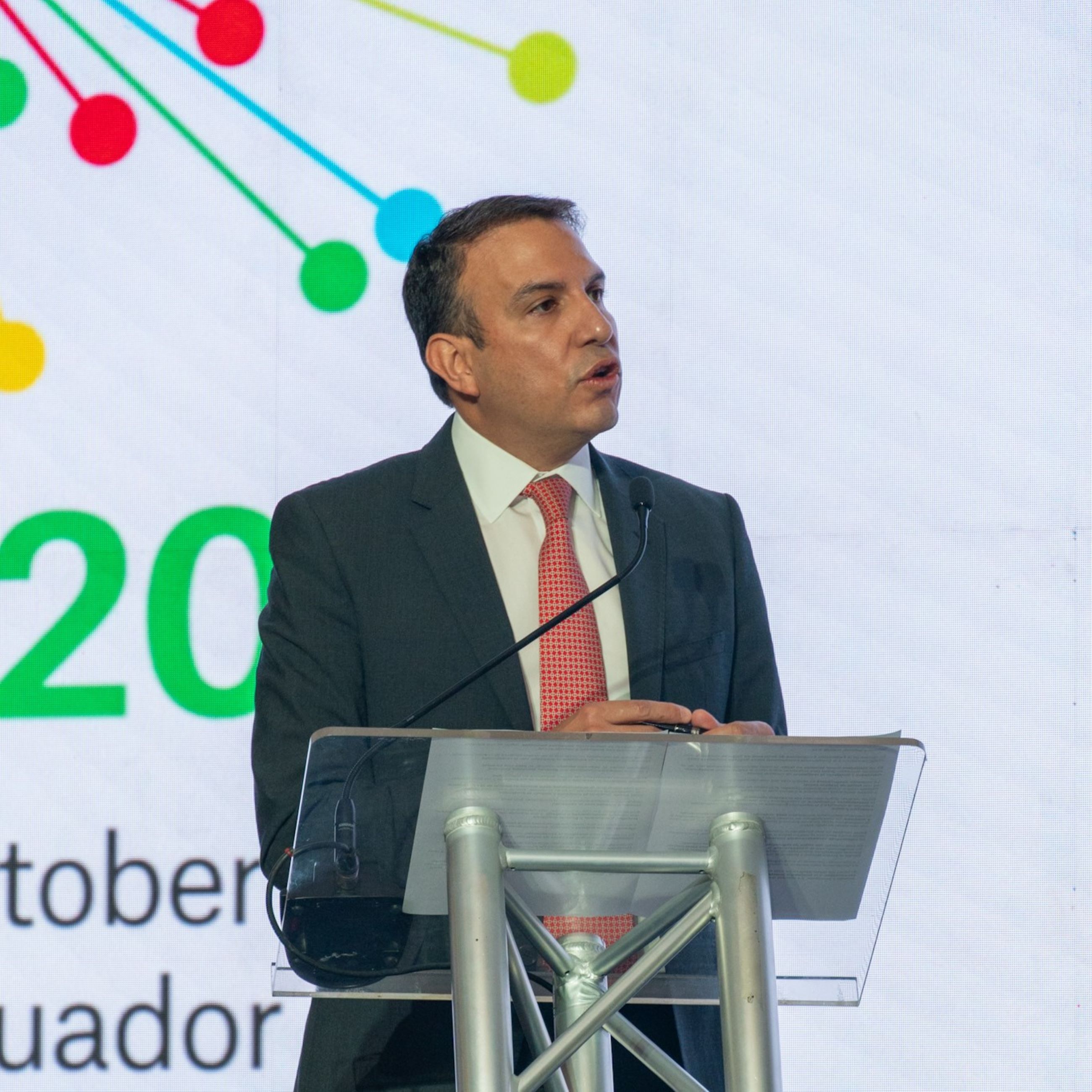 A speaker stands at a podium in front of a colorful backdrop with the text "2025" and "Ecuador".