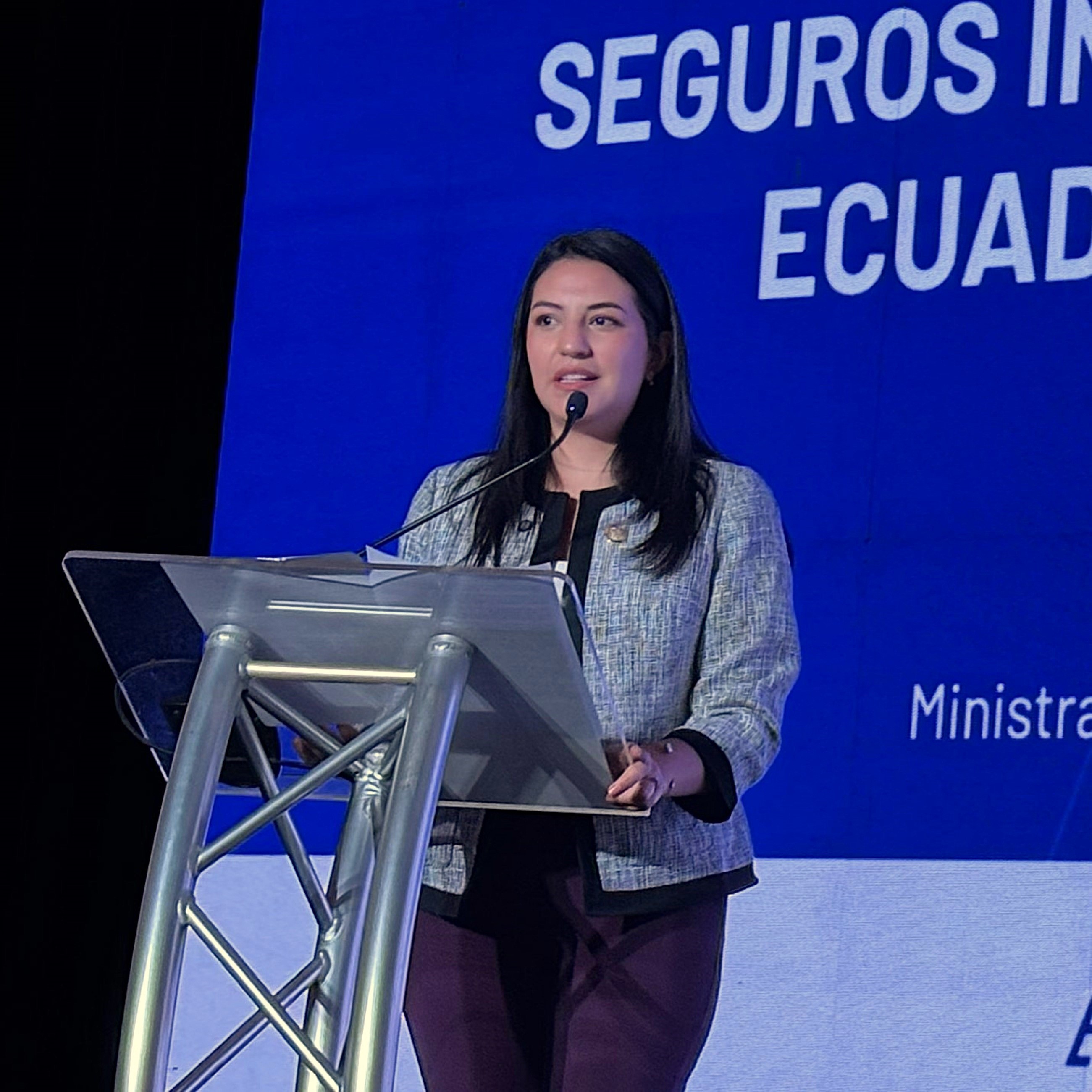 A woman stands at a podium speaking, with a blue background displaying text about insurance in Ecuador.