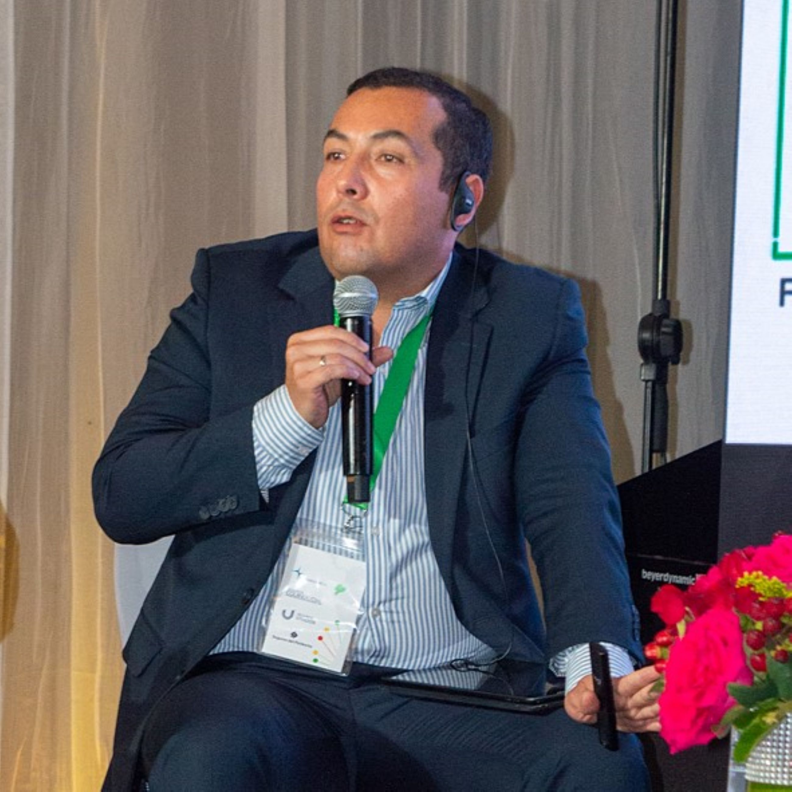 A man in a suit speaks into a microphone while seated next to a woman, with a presentation screen and flowers nearby.