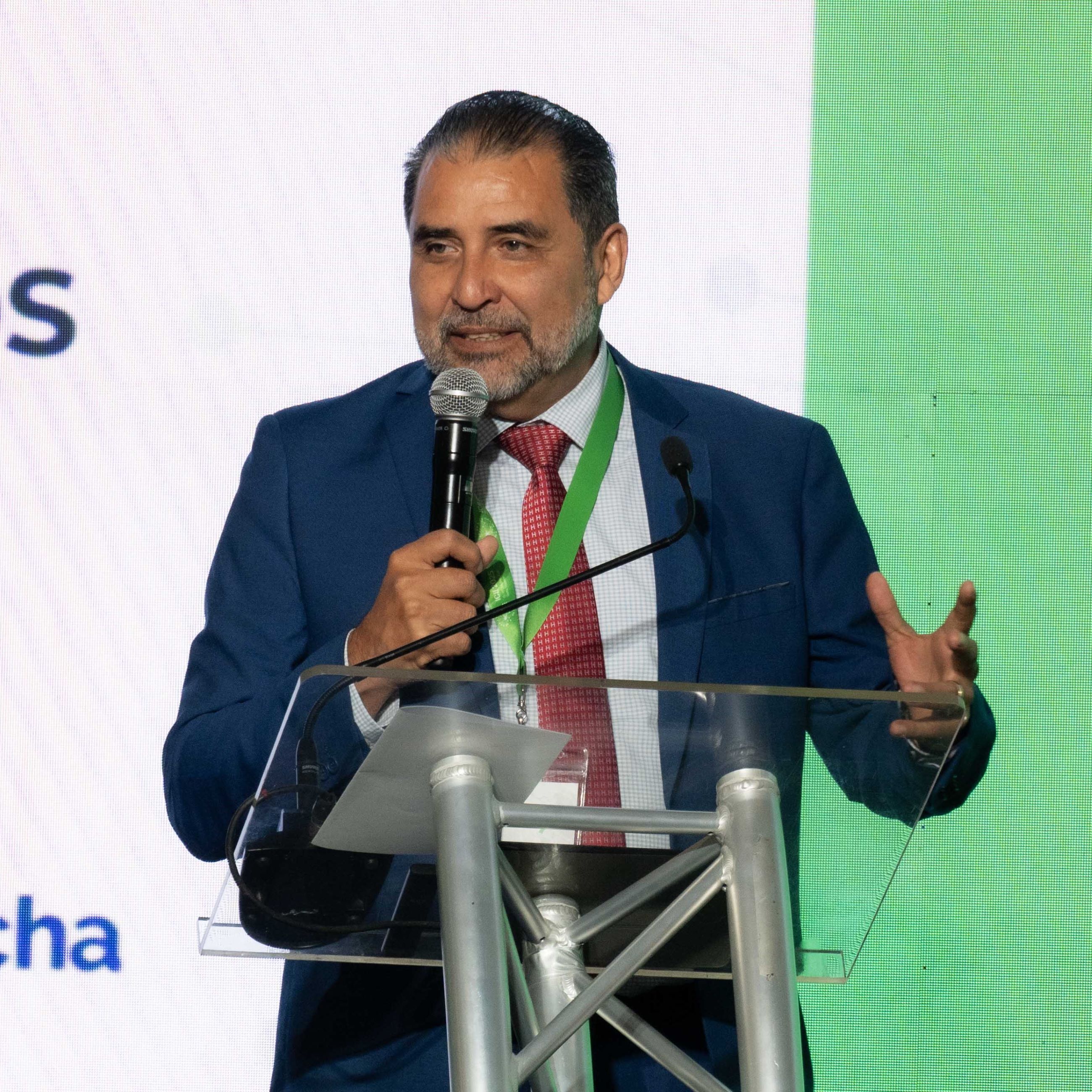 A speaker in a blue suit gestures while holding a microphone at a podium during a presentation.