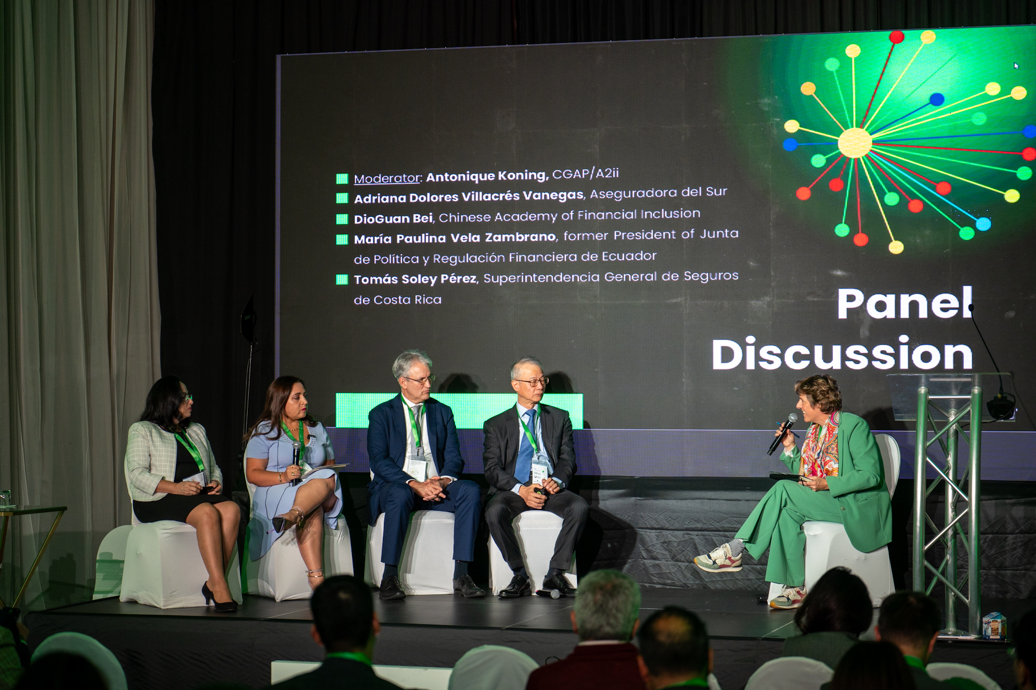 Panel discussion with five speakers seated on stage, a screen displaying names and topics behind them.
