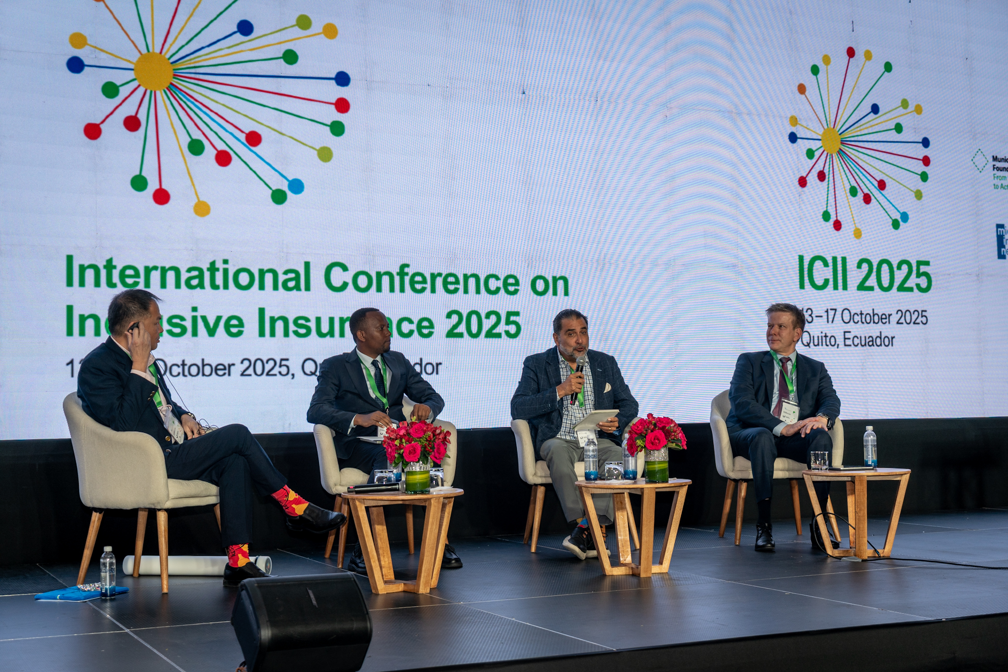 Four speakers sit on stage at a conference, with a backdrop displaying event details and colorful graphics.