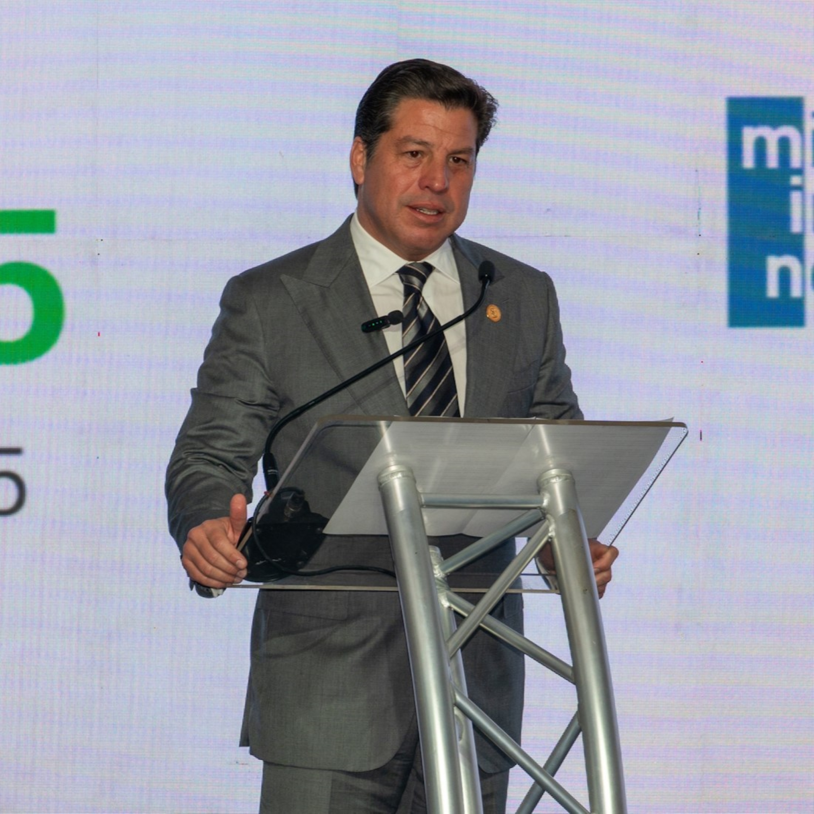 A man in a gray suit stands at a podium, speaking at an event with a large screen behind him.