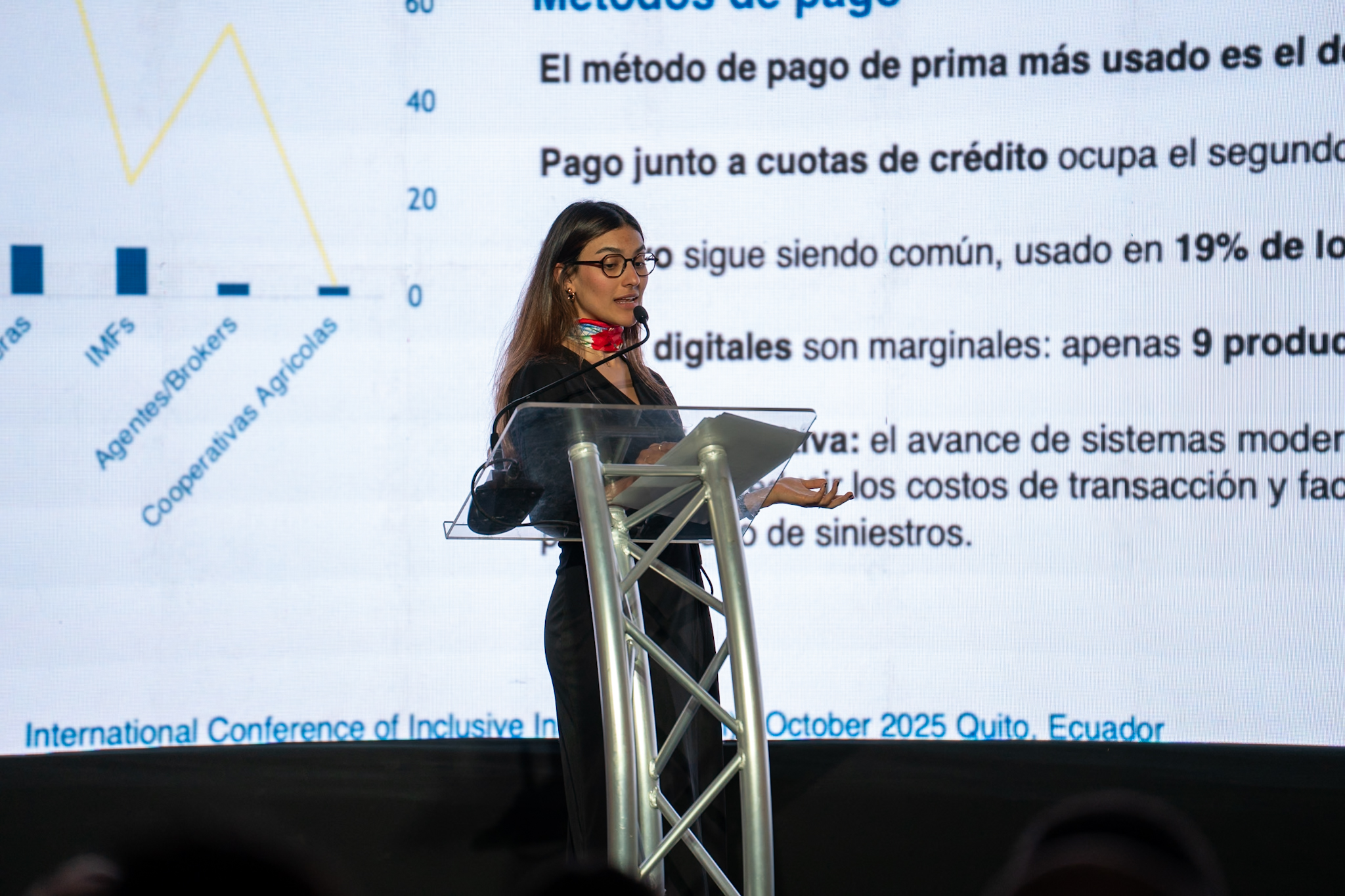 A speaker presents at a conference with a large screen displaying text and graphs in the background.