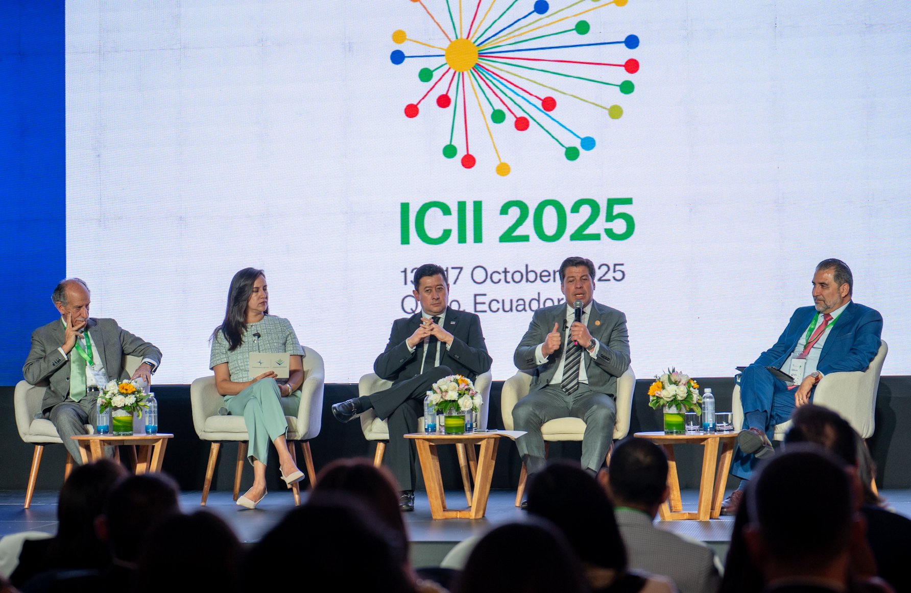 A panel discussion with five speakers on stage, flowers and water bottles on tables, and a large screen behind them.