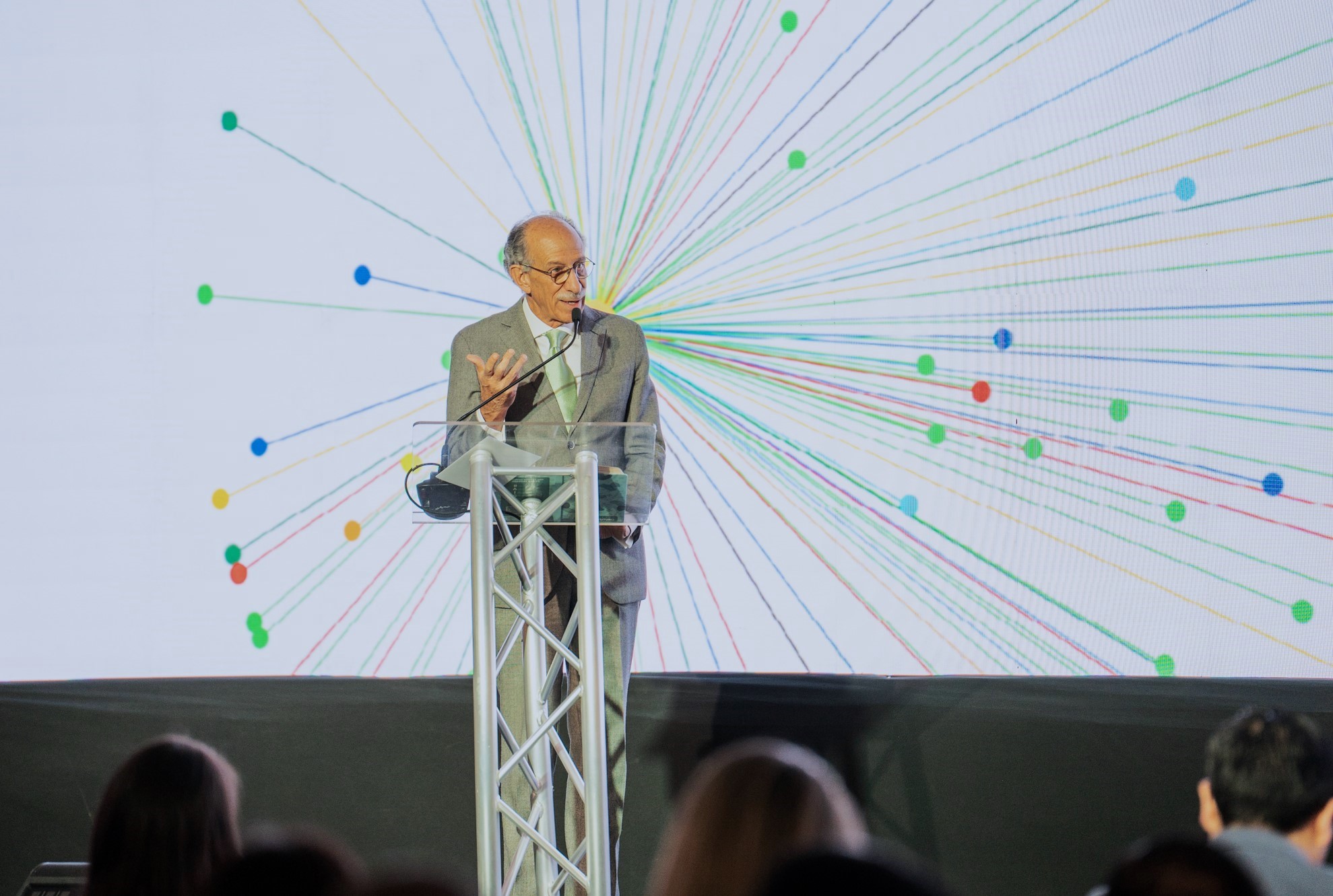 A speaker at a podium gestures, with a colorful radial design projected behind him.
