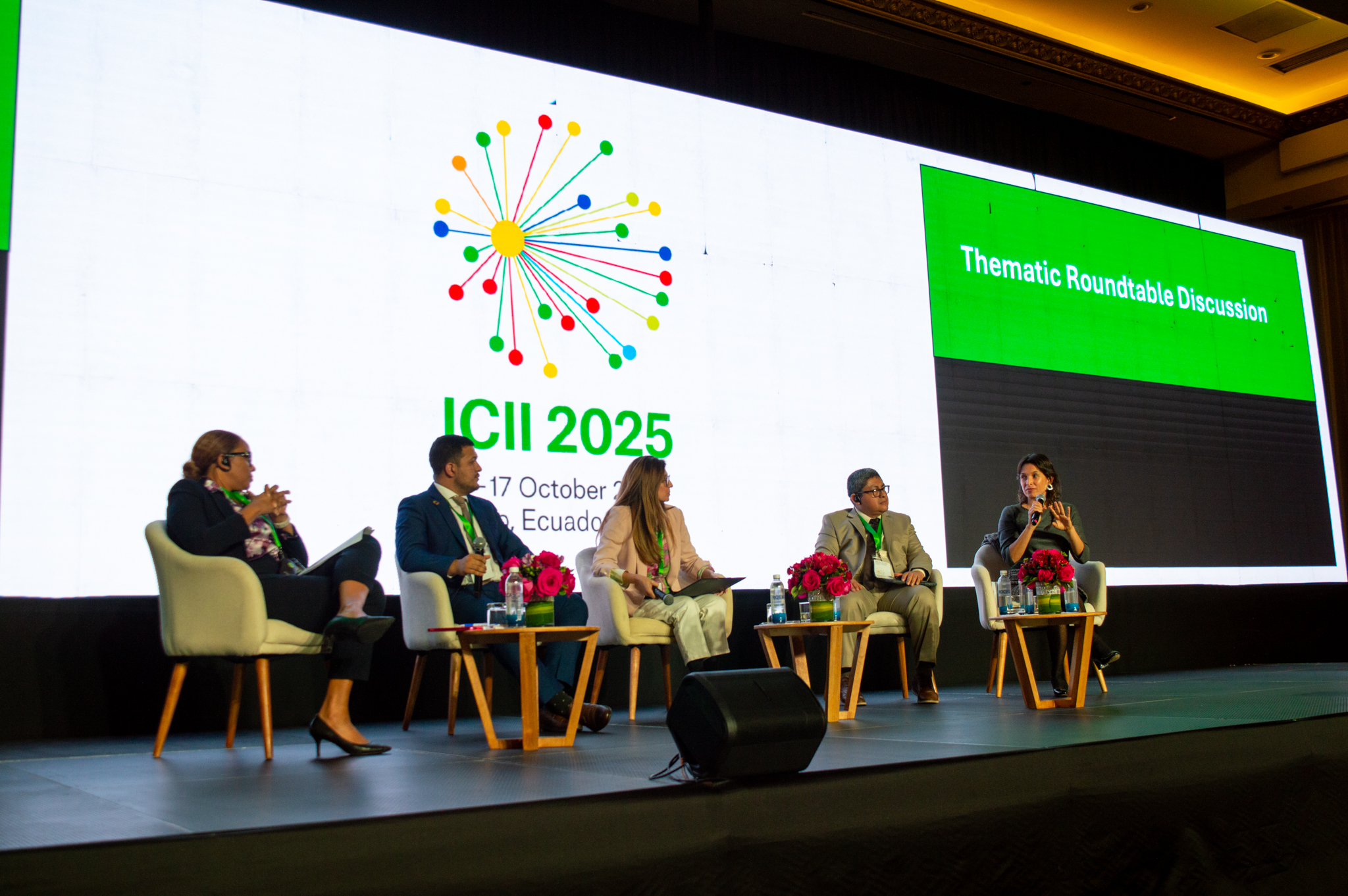 A panel discussion with five speakers on stage, flowers and water bottles on tables, and a large screen behind.