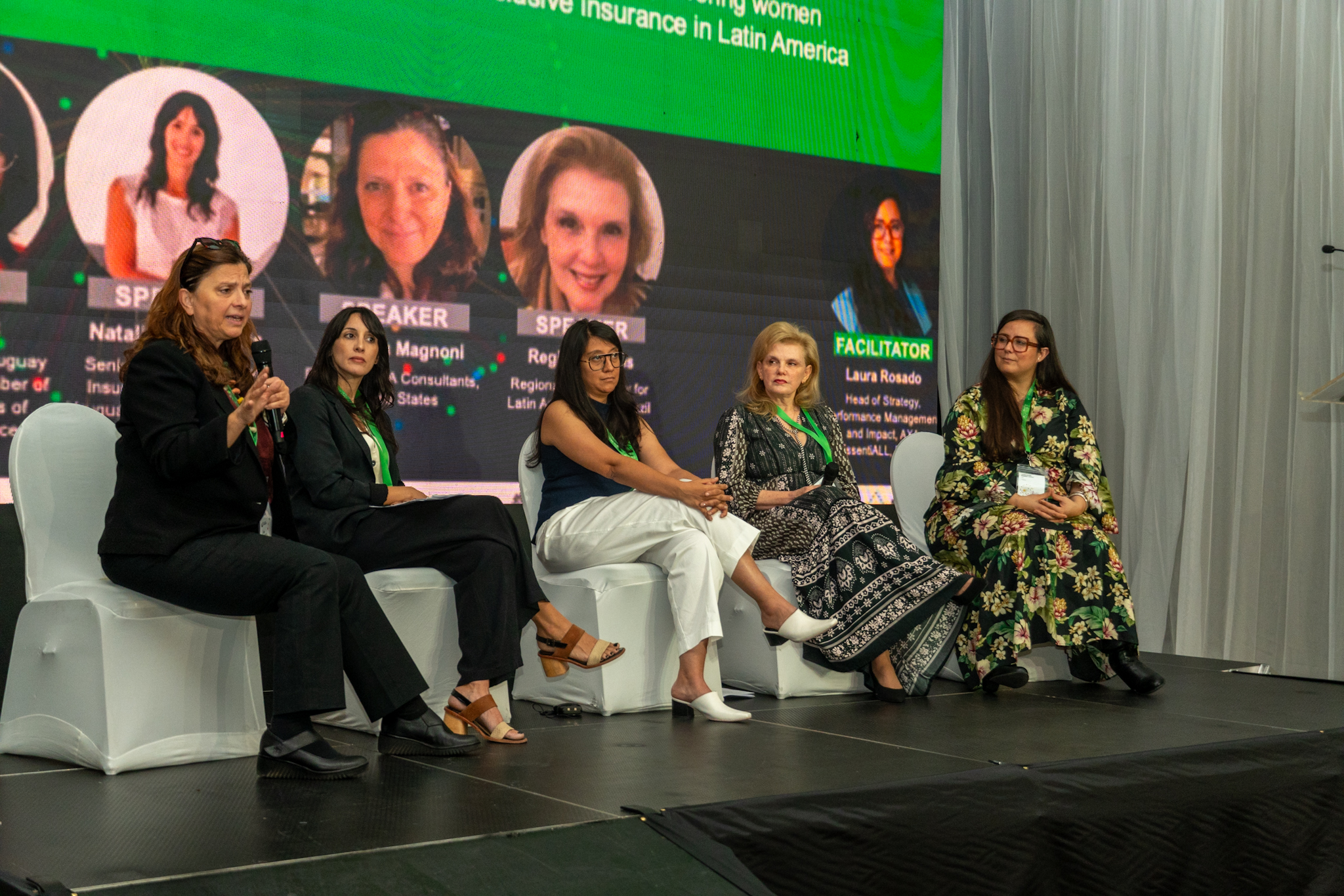 A panel discussion featuring five women seated on stage with a presentation backdrop.