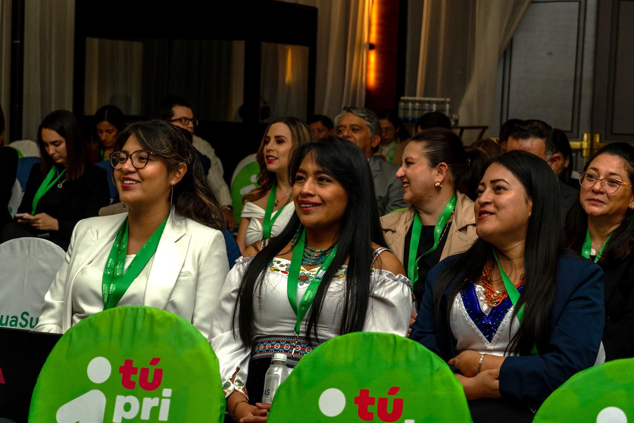 A group of people seated in a conference room, wearing green lanyards and sitting on chairs with green covers.