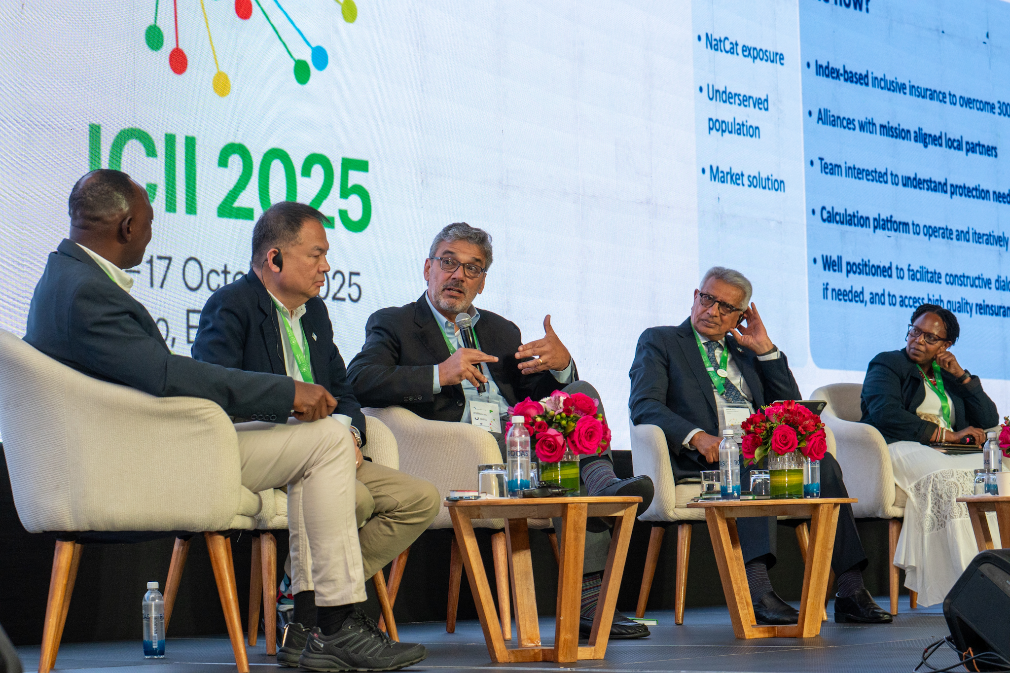 A panel discussion with five speakers seated, flowers and water bottles on tables, and a presentation screen behind.