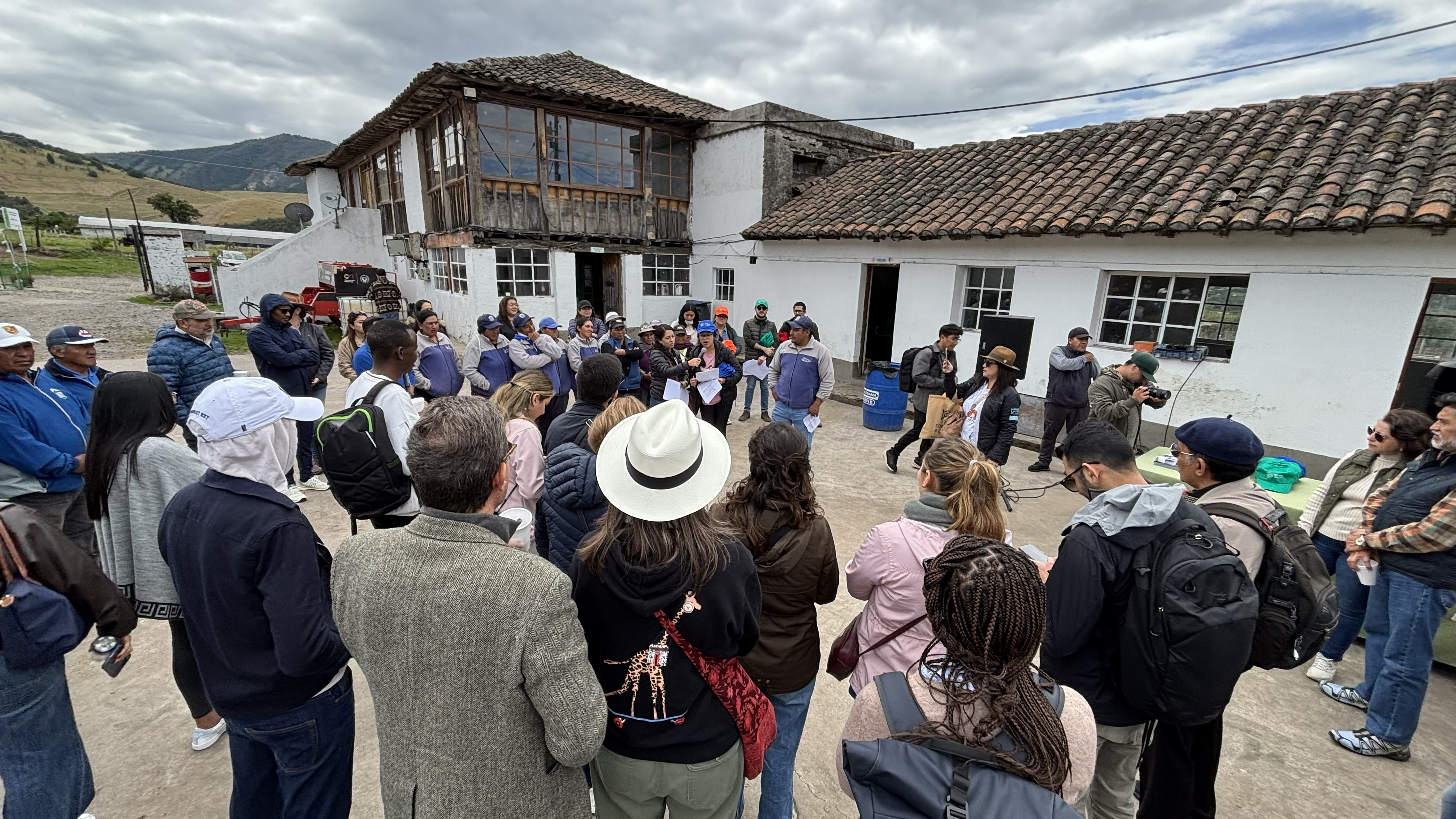 A group of people gathered outdoors near a building, engaged in discussion, with mountains in the background.