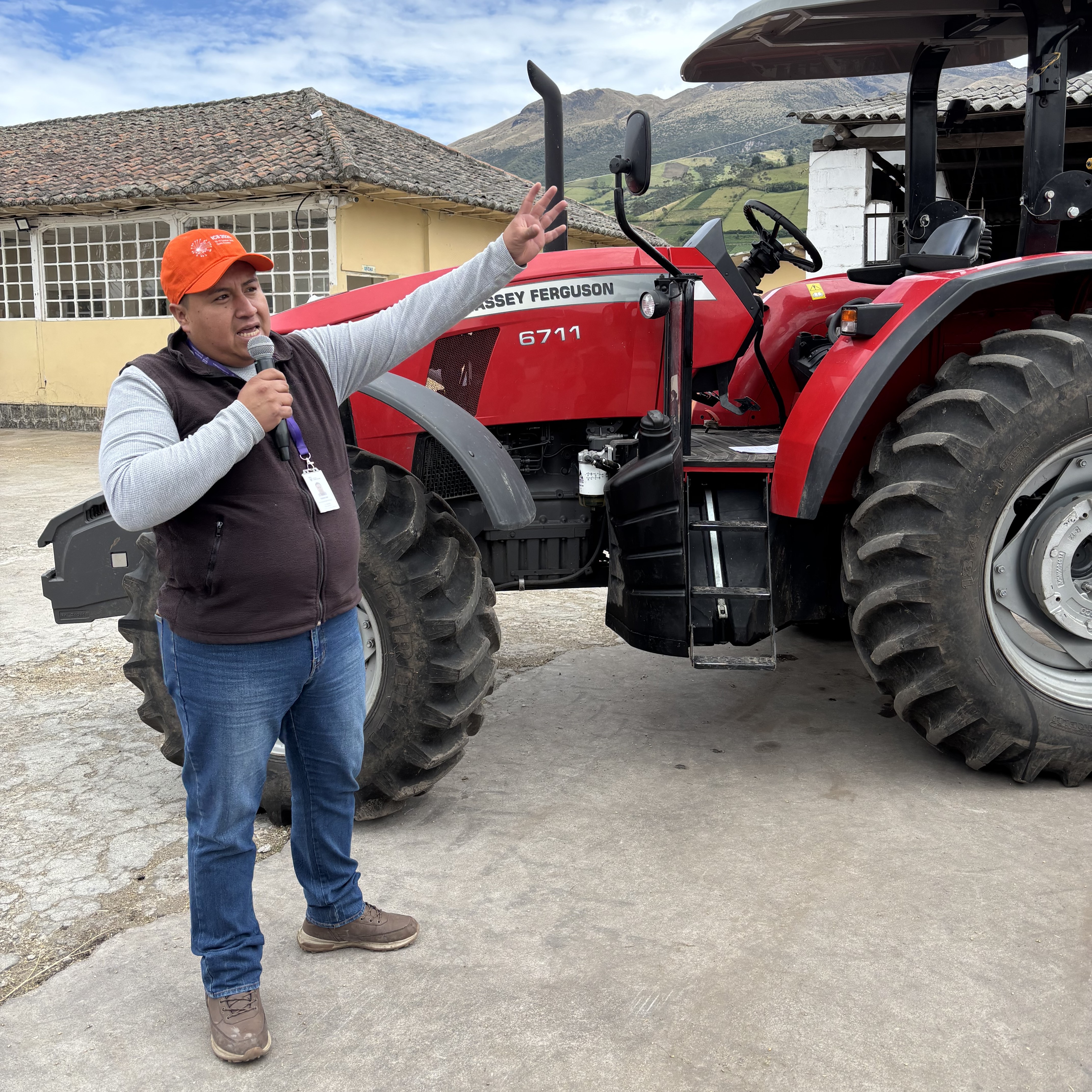 A man stands in a field of green plants, holding a shovel, with mountains and a cloudy sky in the background.
