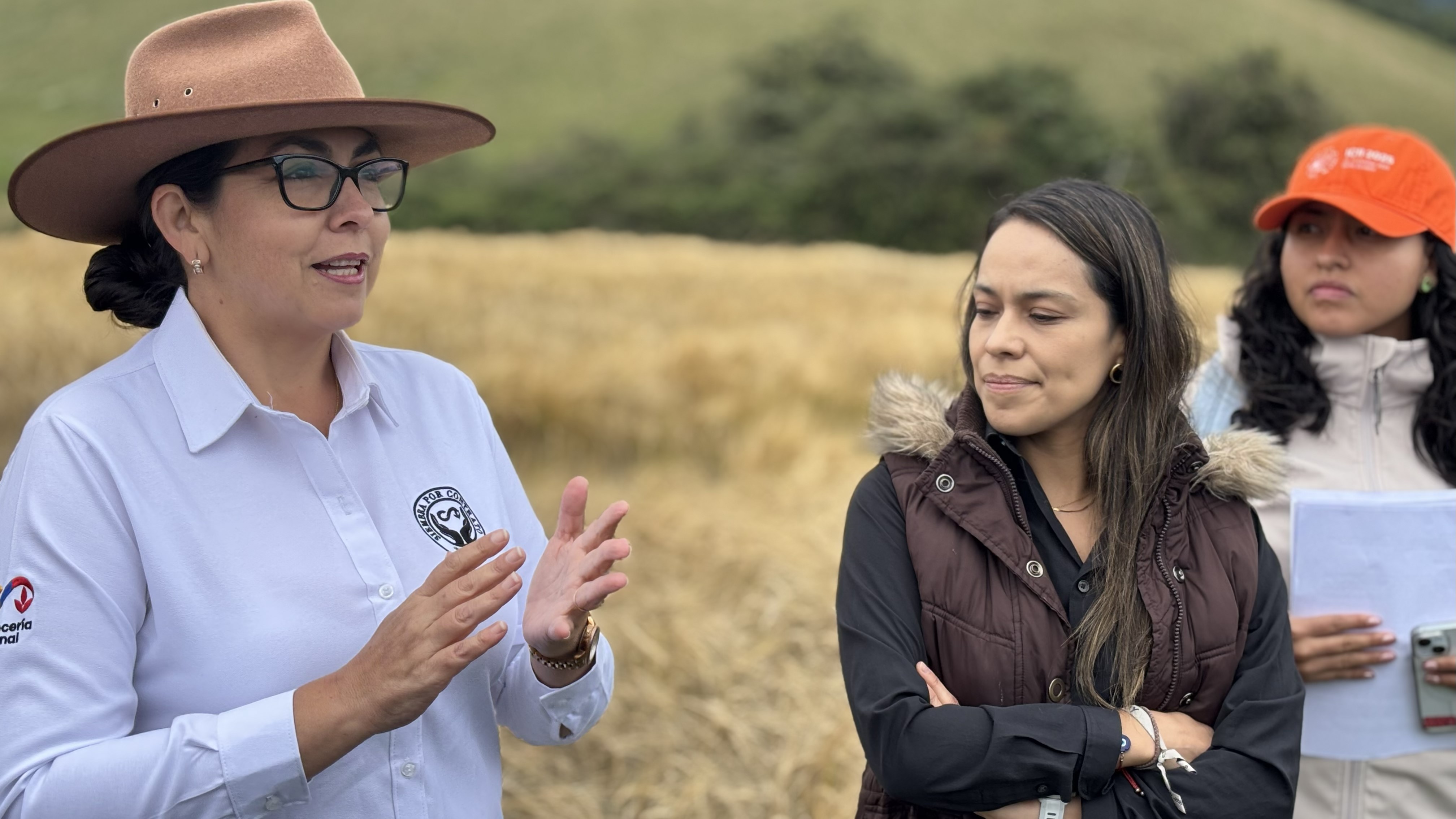 A woman in a white shirt and hat gestures while speaking to two women in a field with golden crops in the background.