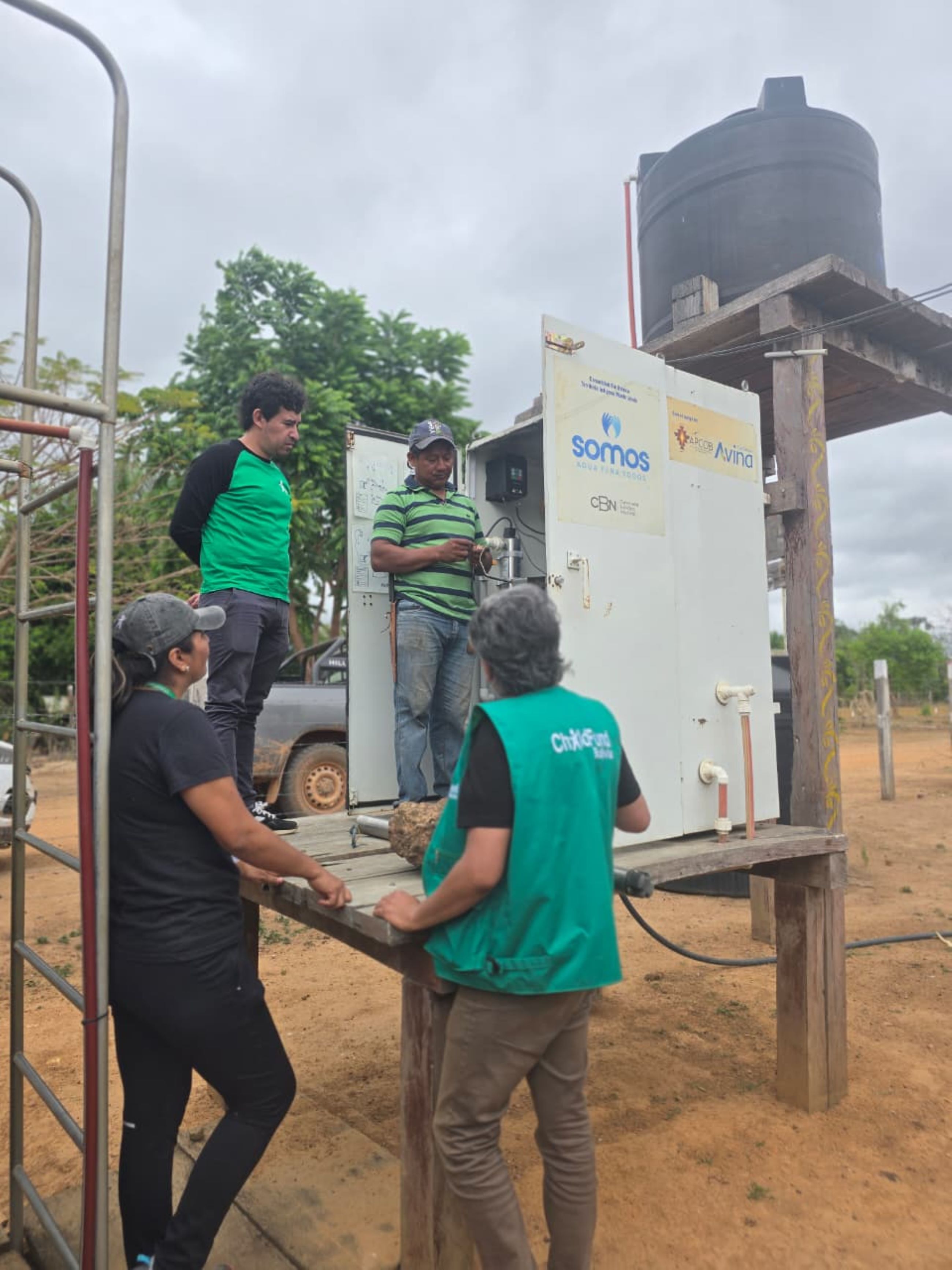 Cuatro personas interactúan con un equipo en un área al aire libre, con un tanque de agua visible en el fondo.