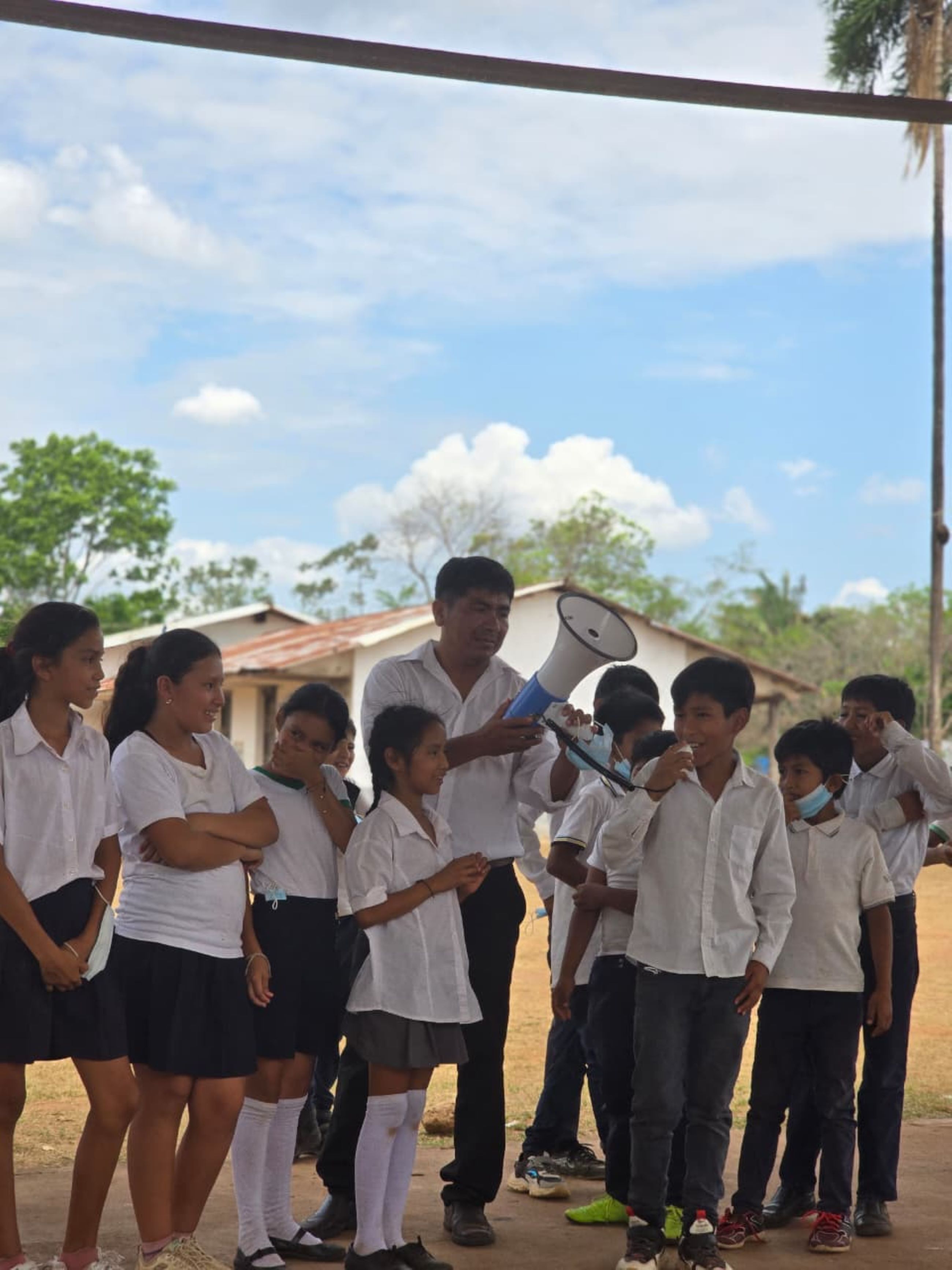 A group of children in white shirts and black skirts stand outdoors, with one person holding a megaphone.