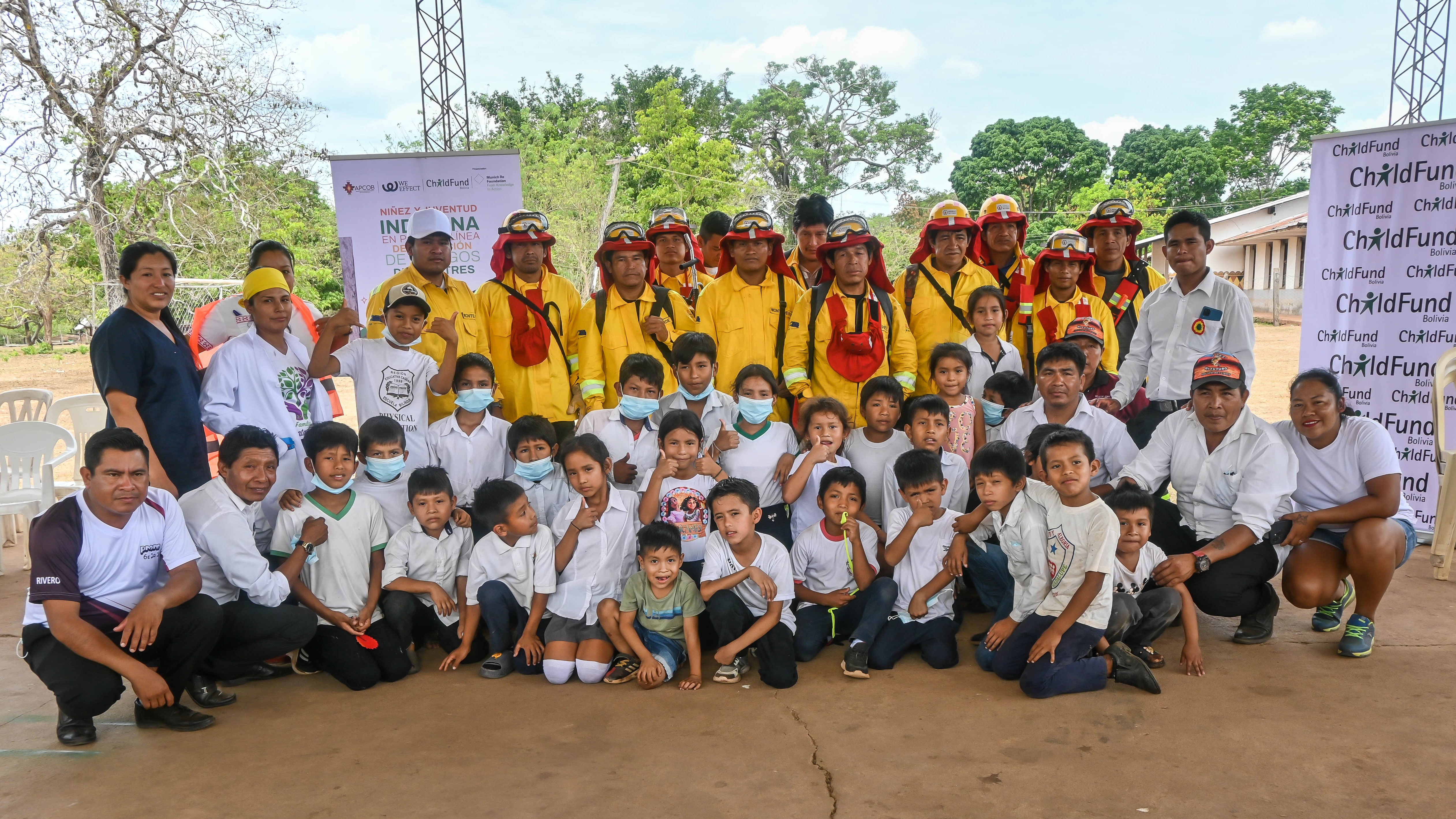 Group photo of children and adults in uniforms, with a banner in the background, outdoors under a clear sky.
