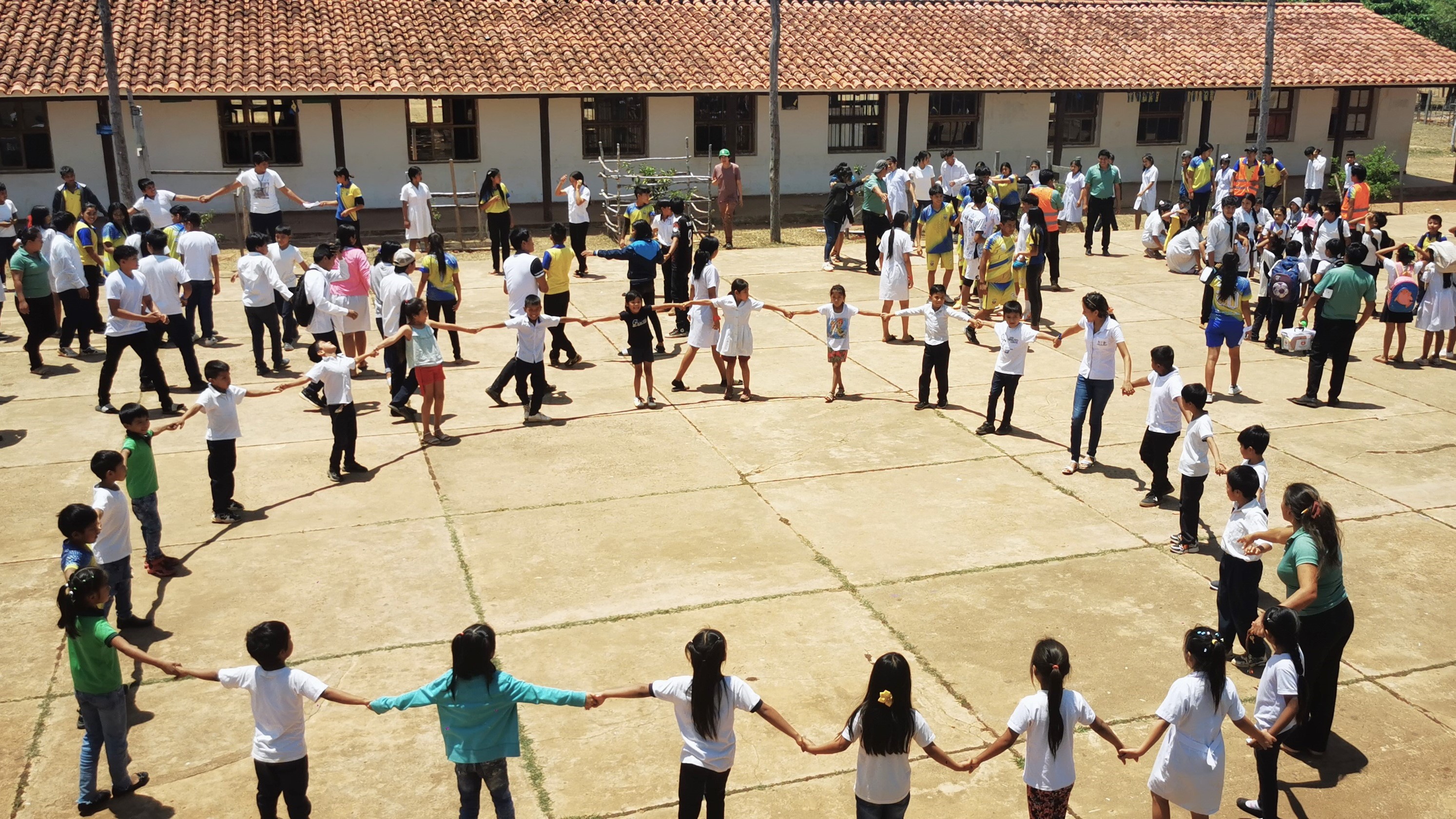 A group of children and adults participate in a circle dance on a sunny day in a schoolyard.