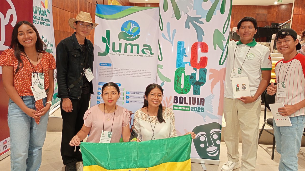 A group of six people poses with a green and yellow flag in front of banners promoting environmental initiatives.
