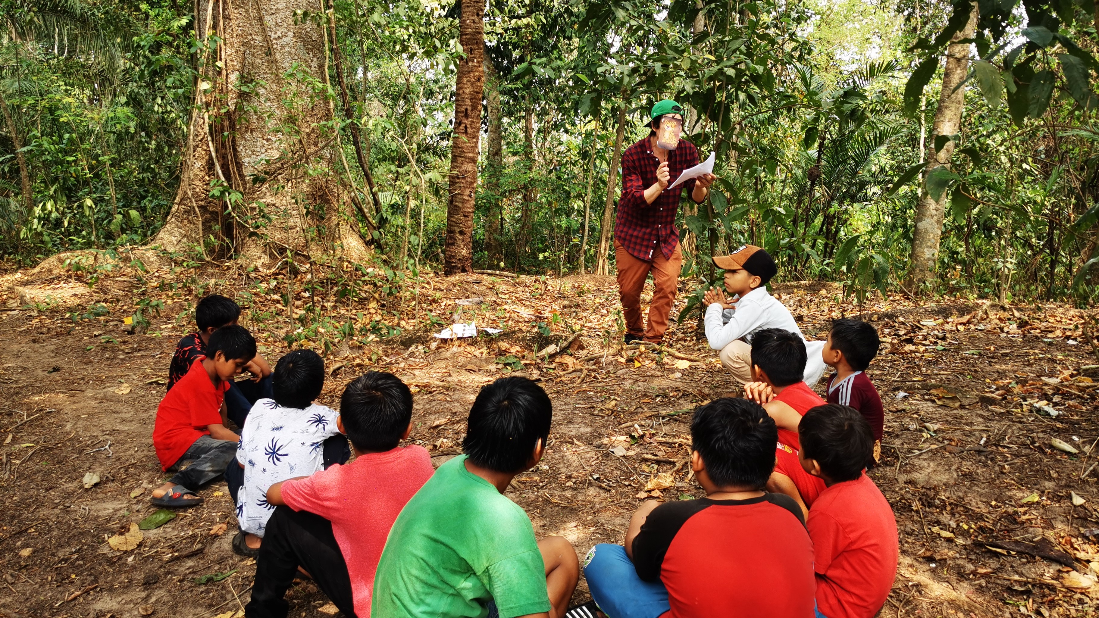 A group of children sitting on the ground, listening to an adult speaking in a forested area.